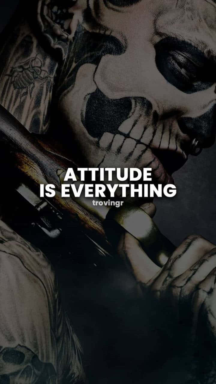語錄 桌布 壁紙 — 一個有刺青、骷髏臉的人手持槍枝，而「attitude is everything」的文字以粗體白色字母出現。