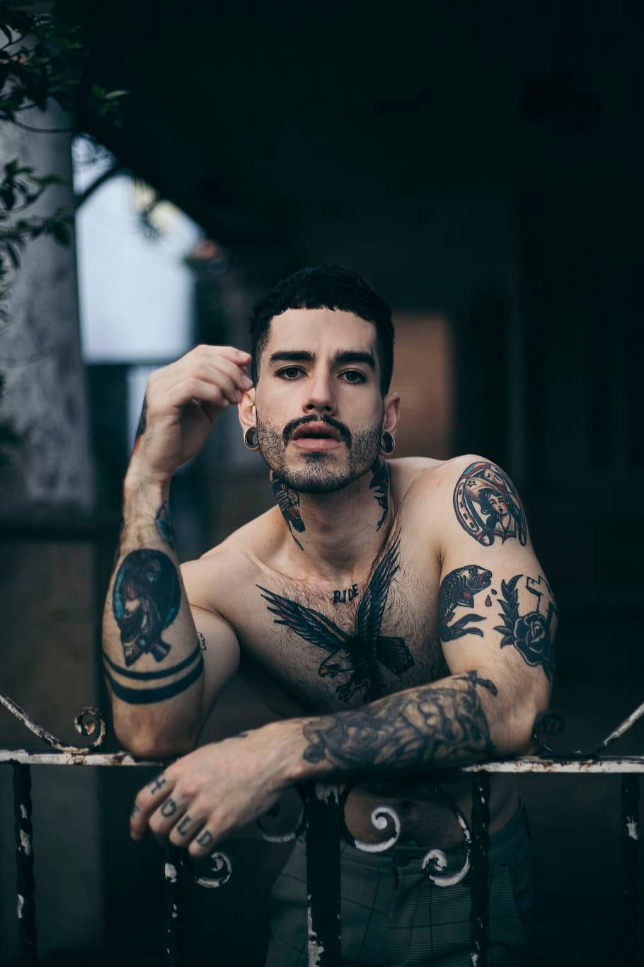 tattoos 桌布 壁紙 — 一個有刺青、鬍鬚和穿環的男人靠在昏暗城市環境中的鍛鐵欄杆上。