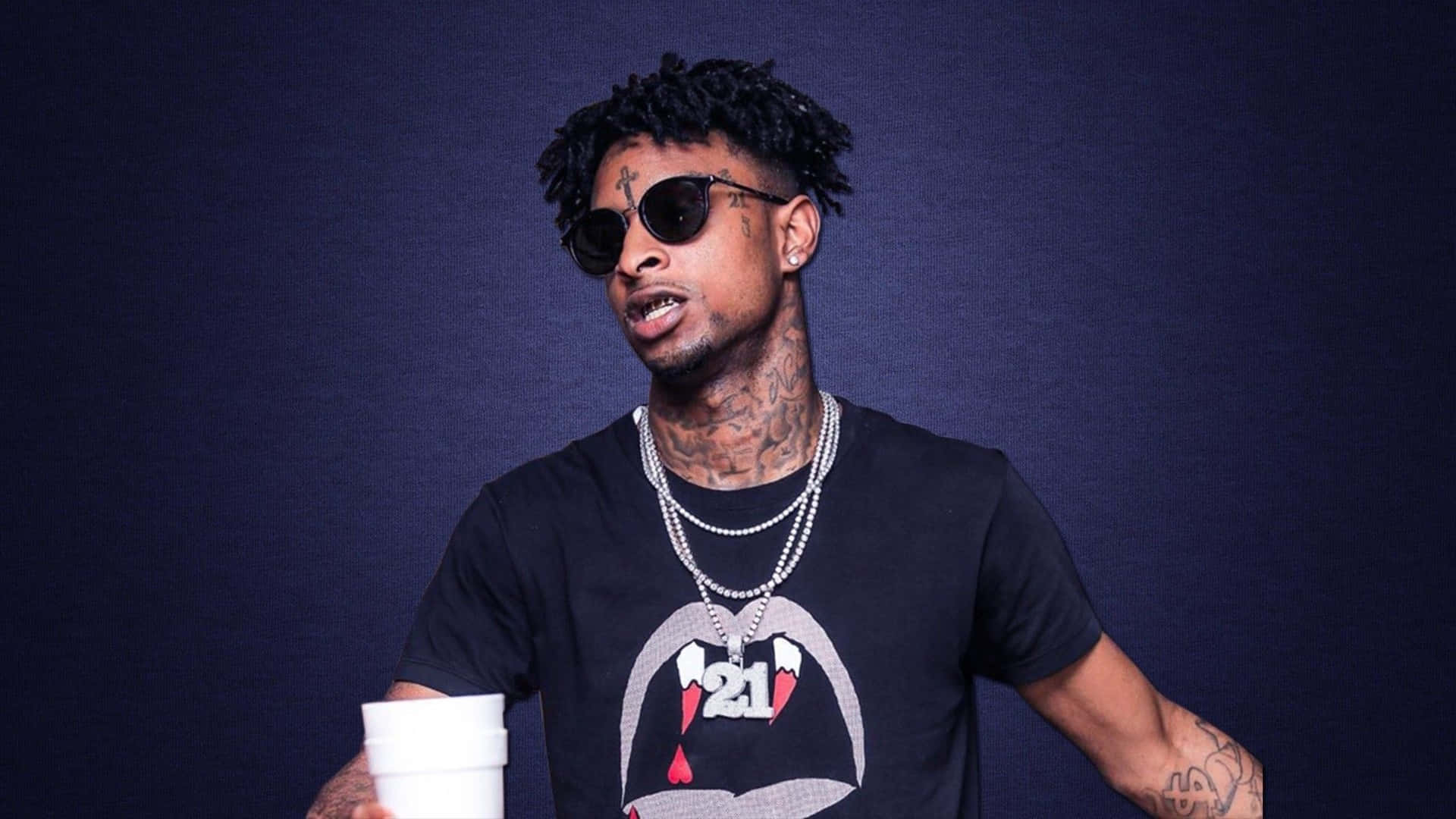 21 Savage 桌布 壁紙 — 一個有刺青和太陽眼鏡的男人在深藍色工作室背景前拿著一個白色杯子。