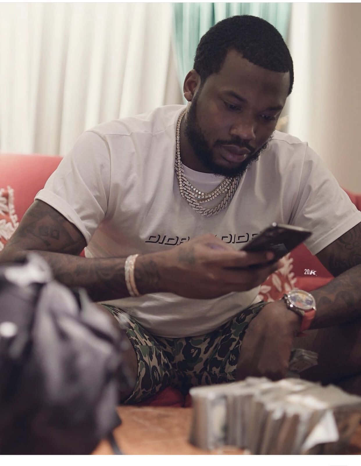Meek 桌布 壁紙 — 一個有刺青和層疊鏈條的男人坐在紅色沙發上，專注於他的手機，附近有成堆的現金。