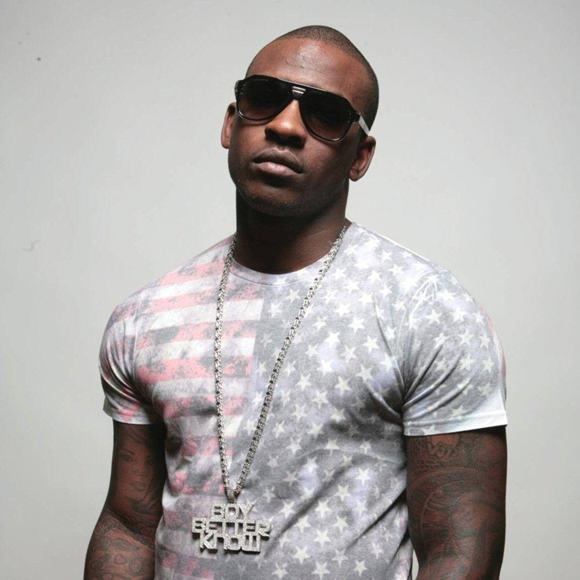 Skepta 桌布 壁紙 — 一個有刺青和戴太陽眼鏡的男人，在純色背景前穿著星條紋T恤和一條「Boy Better Know」鏈條。