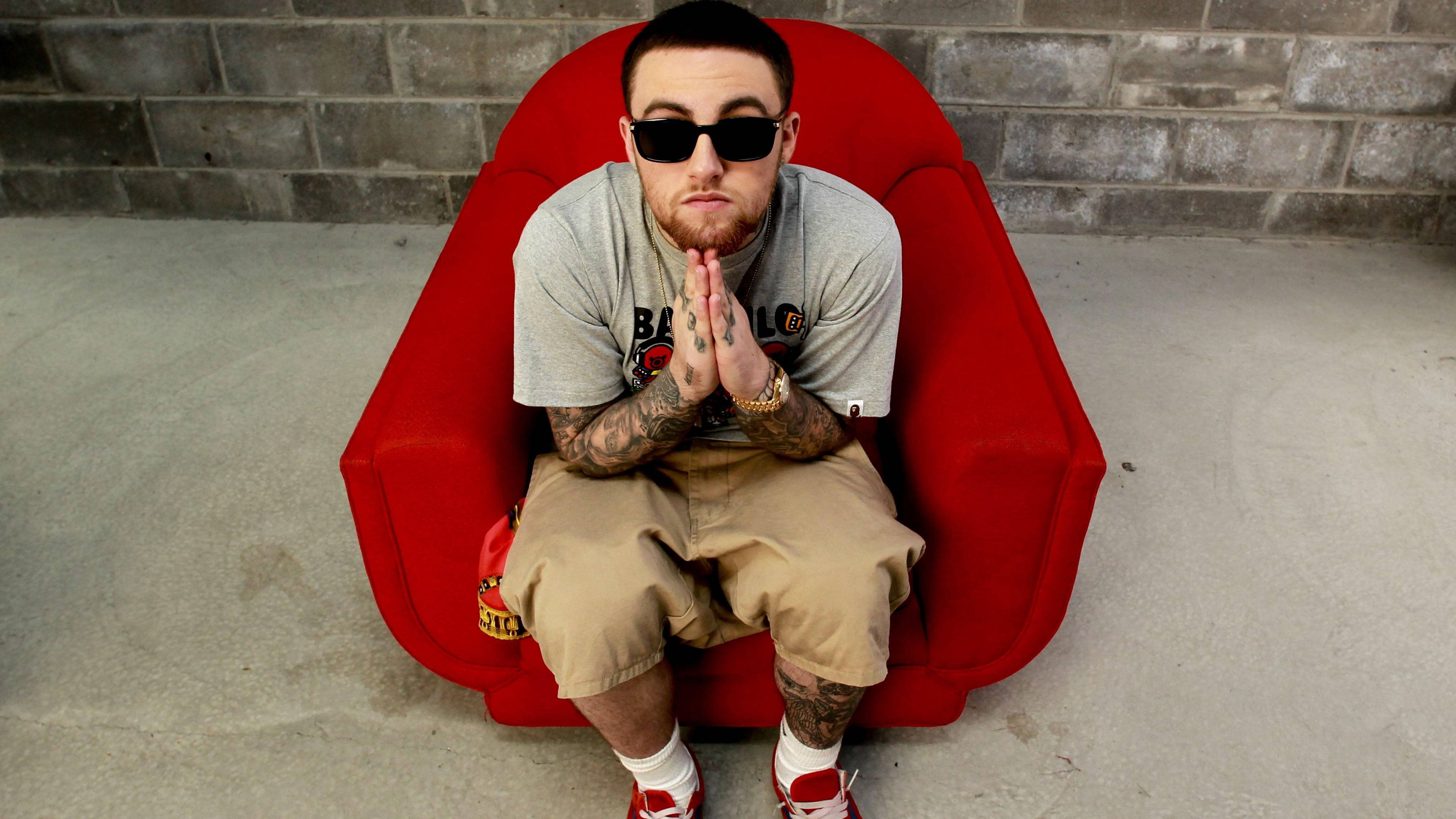 mac miller 4K 桌布 壁紙 — 一個有刺青和戴太陽眼鏡的男人，盤腿坐在一張鮮紅色的扶手椅上，背景是灰色磚牆。