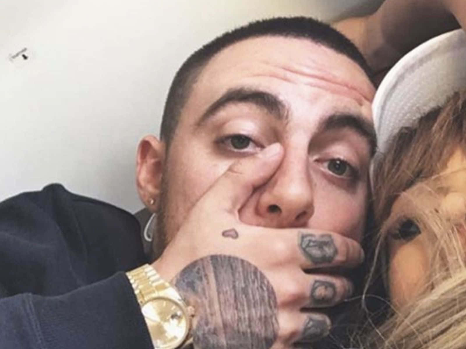 Mac Miller 桌布 壁紙 — 一個有刺青和戴金錶的男人，在擁抱一個戴白帽的女人時摀住自己的嘴。