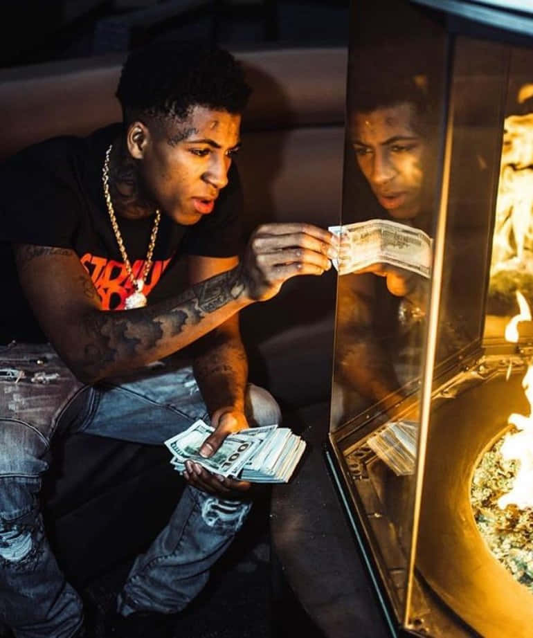 Youngboy 桌布 壁紙 — 一個有刺青和戴金項鍊的男人在壁爐裡燒錢，同時專注地盯著自己的倒影。