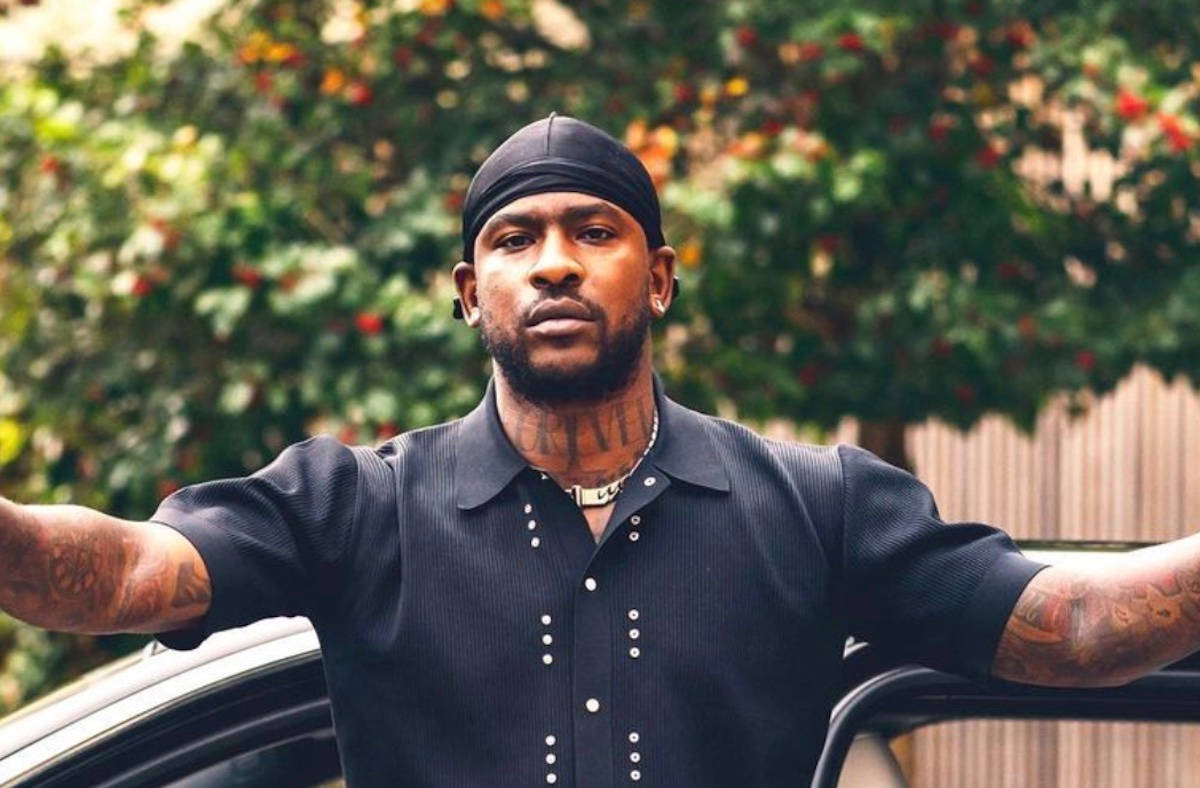 Skepta 桌布 壁紙 — 一個有刺青和戴黑色頭巾的男人自信地靠在一輛車上，雙臂伸展，背景是模糊的綠色樹葉。