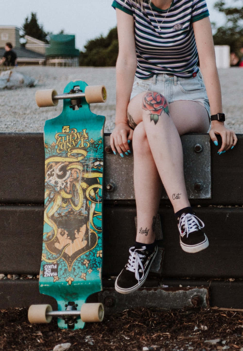 Retro Skateboard 桌布 壁紙 — 一個有刺青和藍色指甲的人坐在金屬長椅上，旁邊放著一塊色彩繽紛的長板，在黃昏時分。