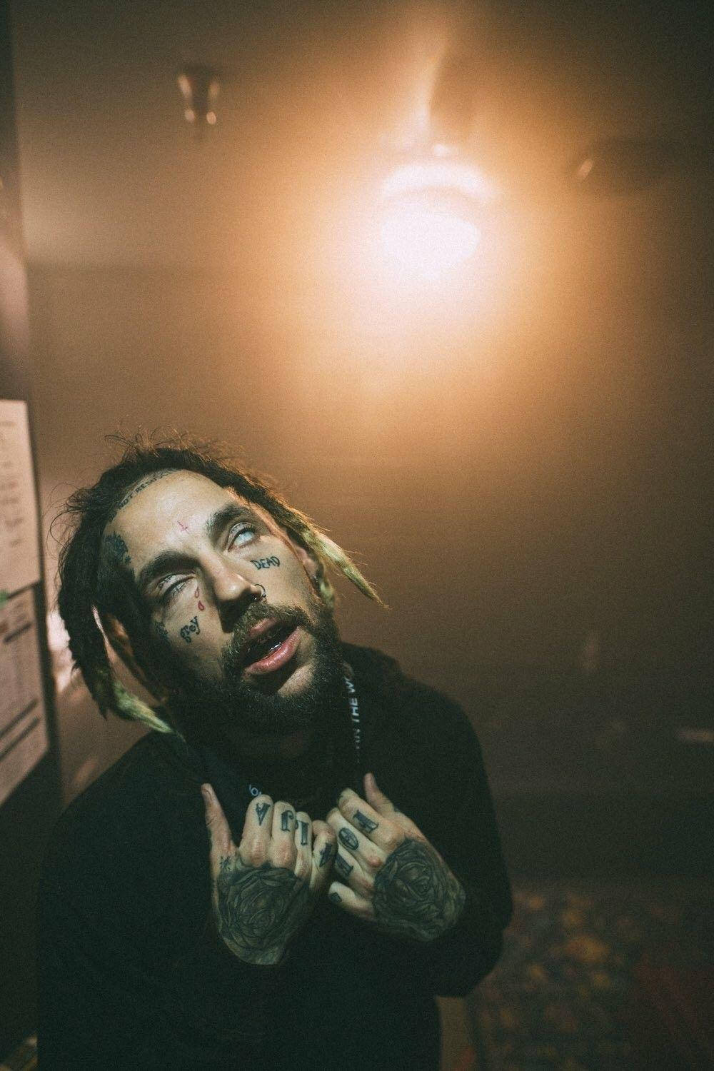 suicideboys 桌布 壁紙 — 一個有刺青和辮子的男人在昏暗煙霧瀰漫的房間裡，抬頭仰望一盞溫暖發光的燈泡。