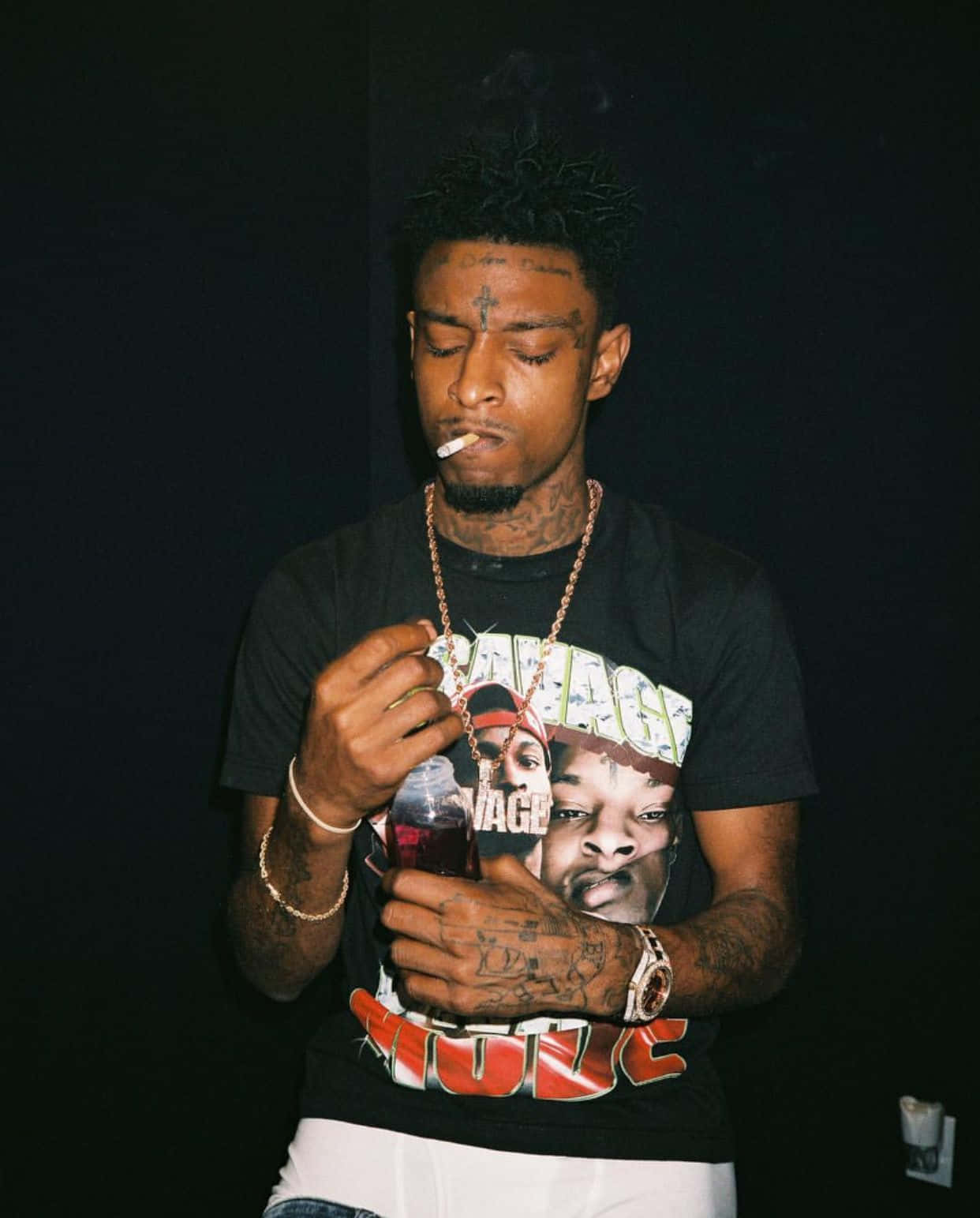 21 Savage 桌布 壁紙 — 一個有刺青和額頭上有十字架的男人，在黑暗背景下抽煙並拿著飲料。
