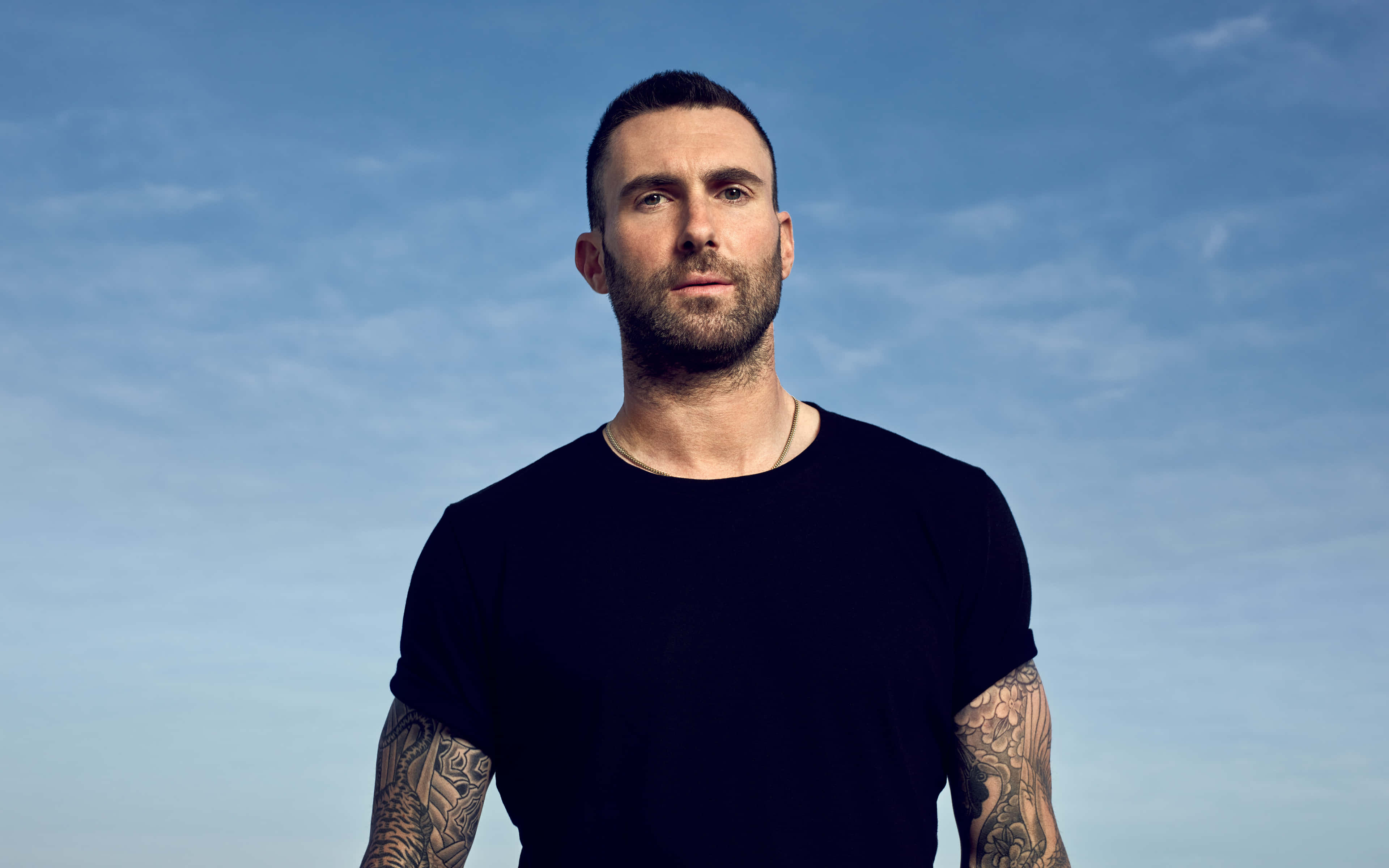 Adam Levine 桌布 壁紙 — 一個有刺青和鬍鬚的男人在晴朗的藍天下直視鏡頭。