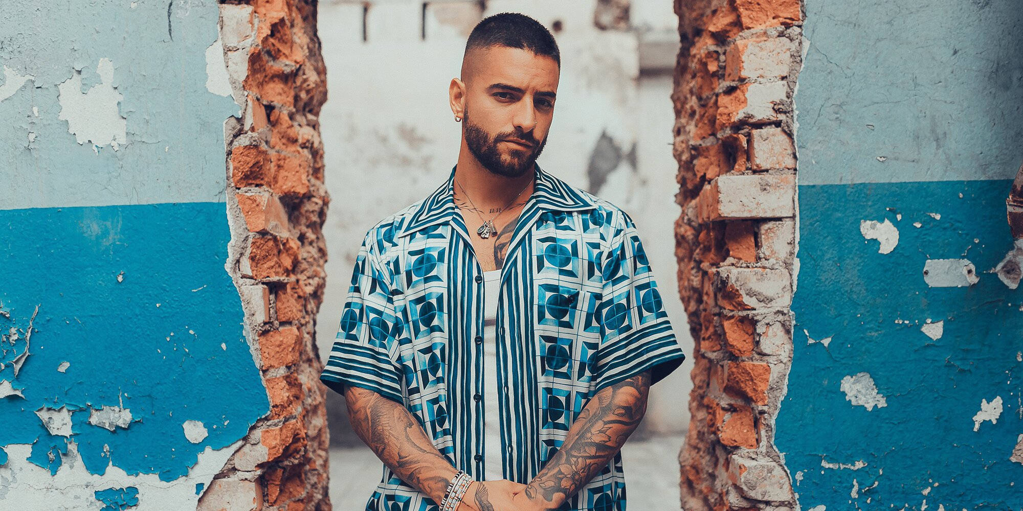 Maluma 桌布 壁紙 — 一個有刺青和鬍鬚的男人站在破爛的磚牆之間，背景是風化的藍白牆壁。