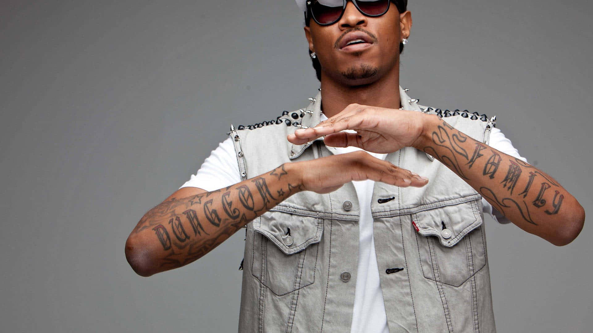 Rappers 桌布 壁紙 — 一個有刺青手臂和戴太陽眼鏡的男人在純灰色背景前用手做出心形。