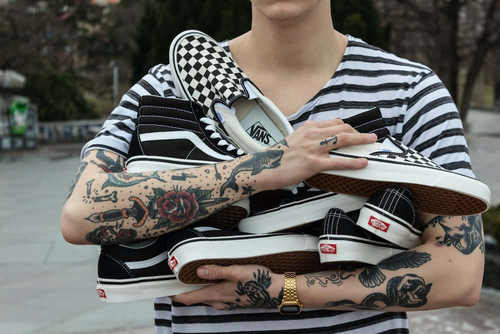 Vans 桌布 壁紙 — 一個有刺青手臂的人，在模糊的戶外背景下，拿著一堆黑白Vans運動鞋。