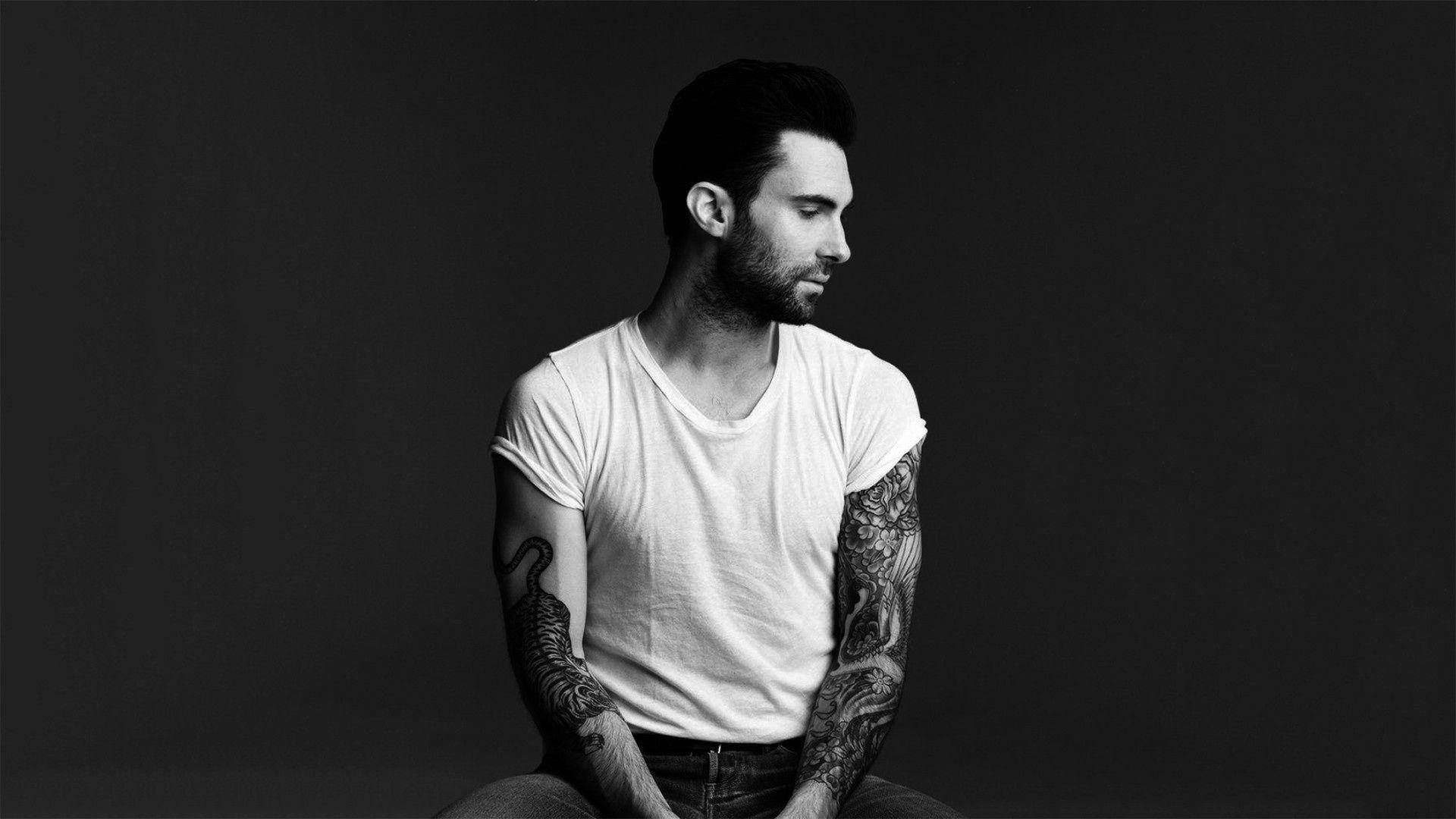 Adam Levine 桌布 壁紙 — 一個有刺青手臂的男人側面坐在深色背景前，穿著白色T恤。