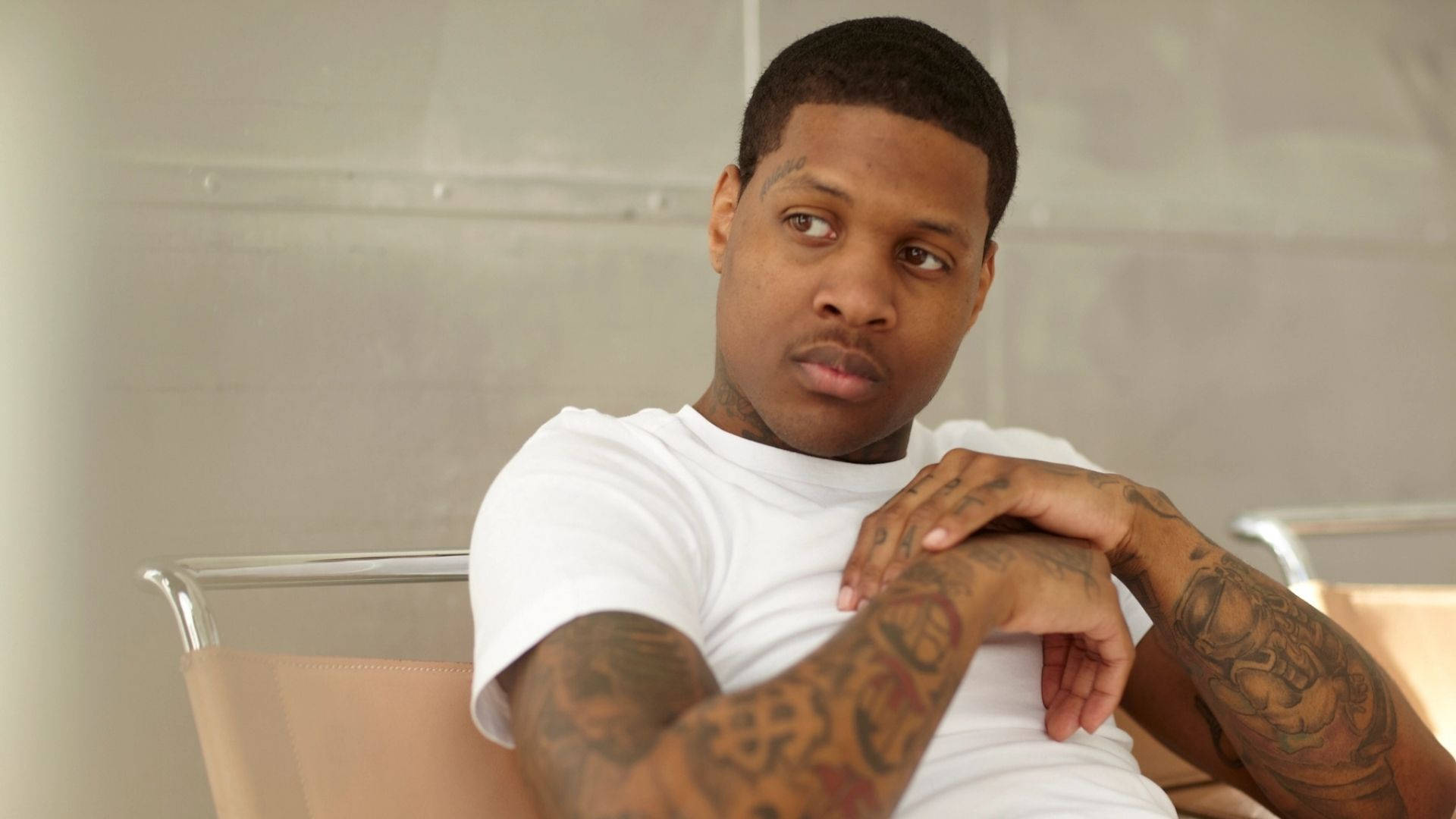 Lil Durk 桌布 壁紙 — 一個有刺青手臂的男人坐在椅子上，穿著白色T恤，嚴肅地望向一邊。