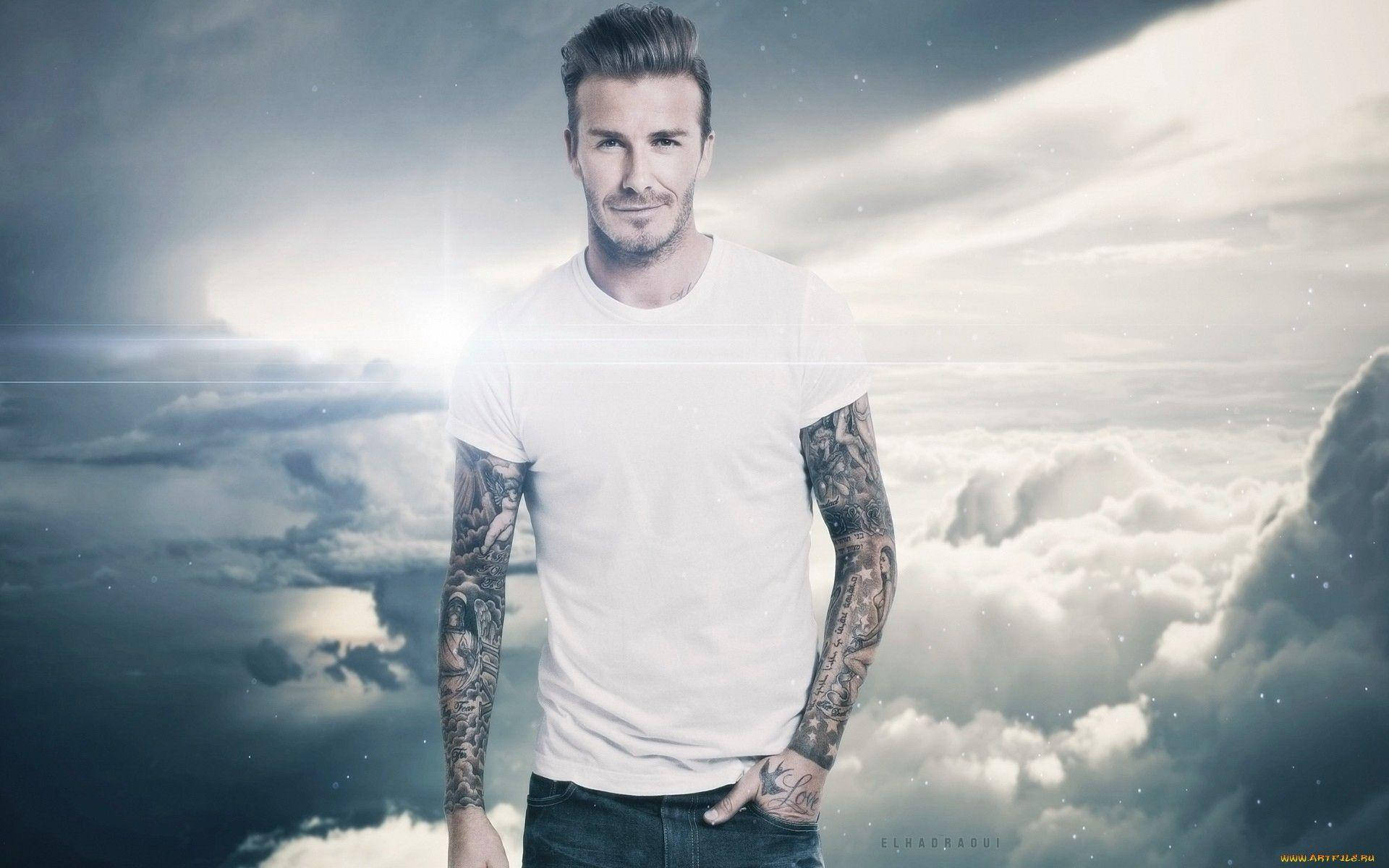 David Beckham 桌布 壁紙 — 一個有刺青手臂的男人自信地穿著白色T恤，站在戲劇性的多雲天空和明亮的陽光下。