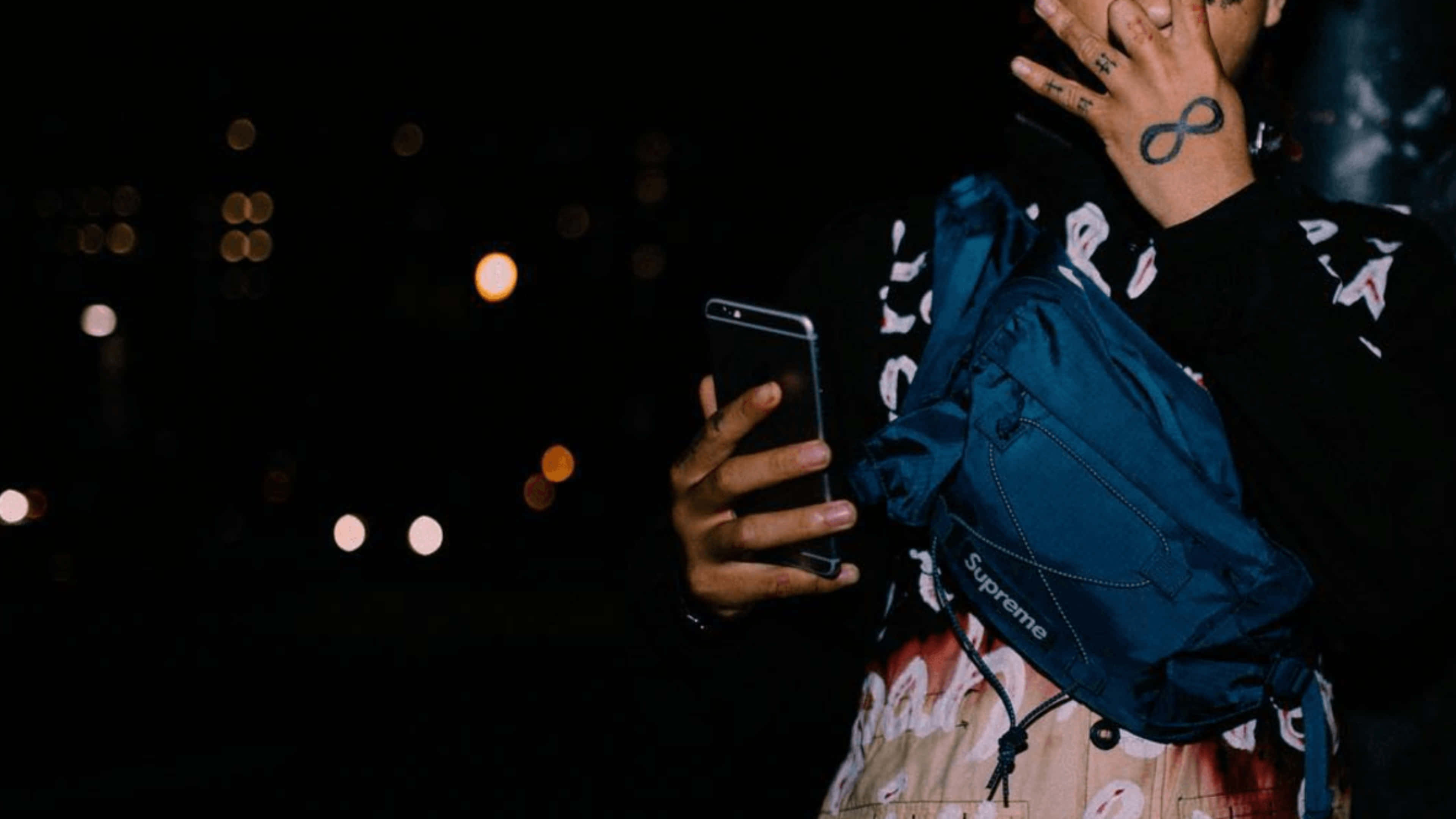 Trippie Redd 桌布 壁紙 — 一個有刺青的人，在模糊的夜景和發光的散景燈光背景下，拿著一支手機和一個藍色Supreme包包。