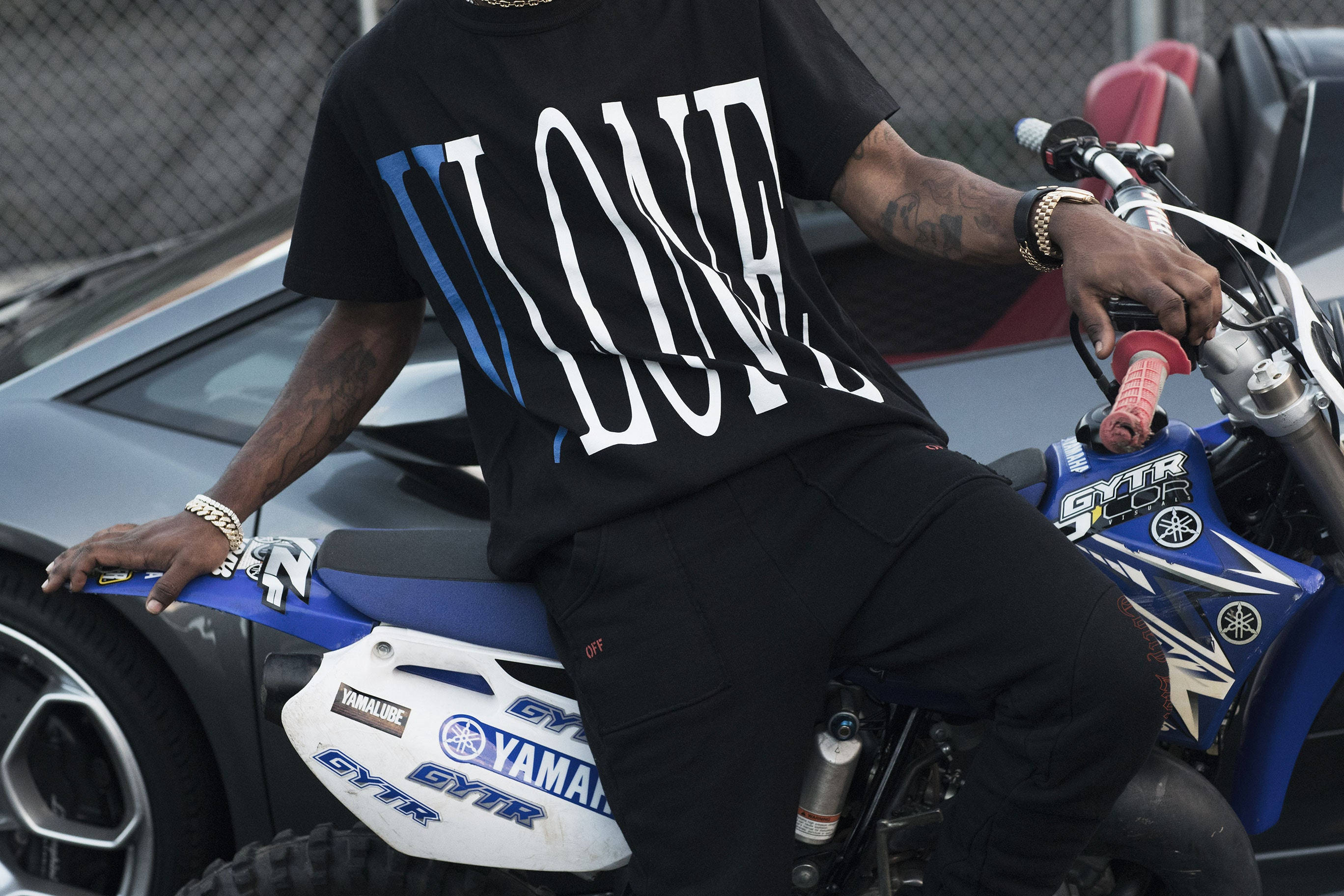 Vlone 桌布 壁紙 — 一個有刺青的人坐在藍白相間的Yamaha越野機車旁邊，旁邊是一輛時尚的灰色跑車。