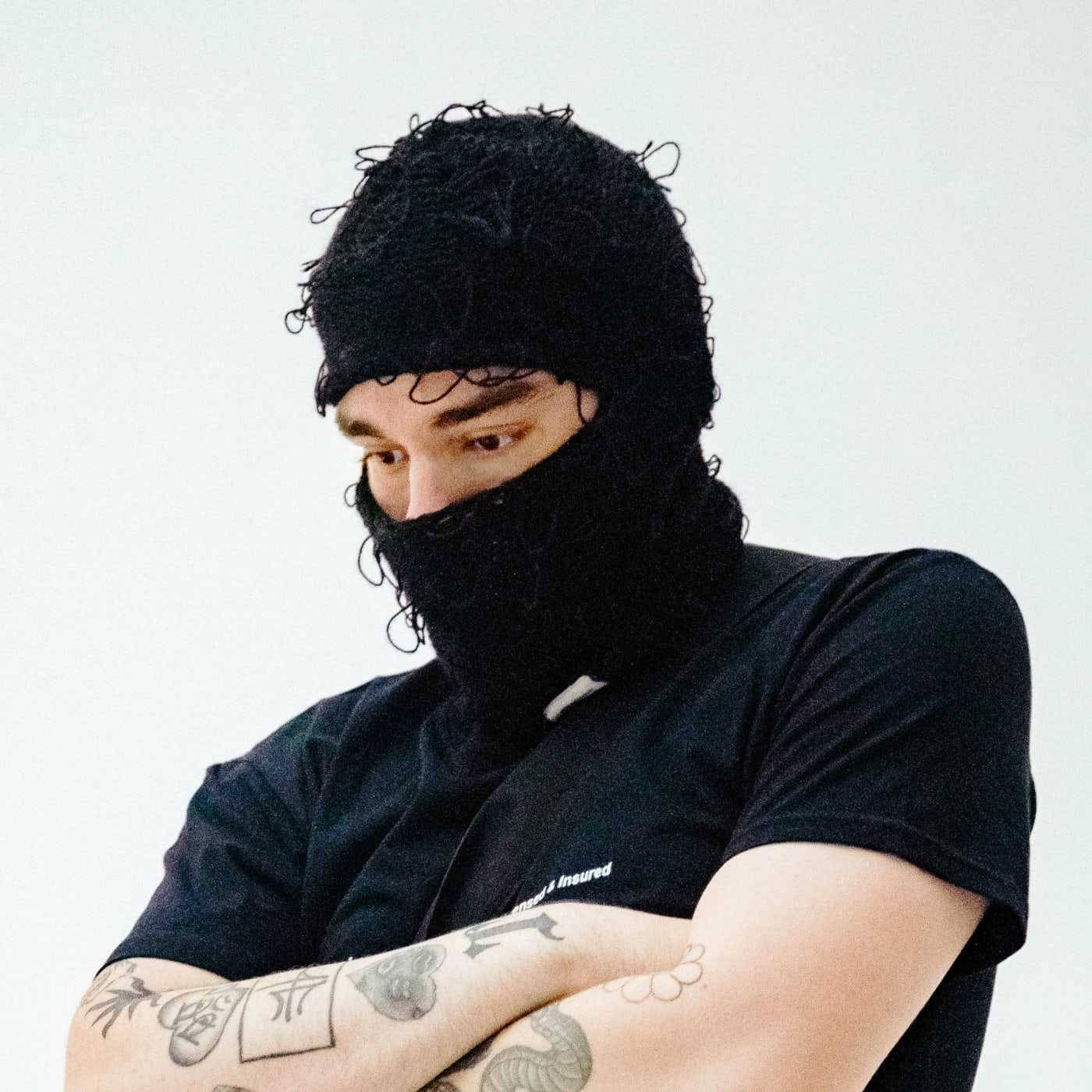說唱歌手 桌布 壁紙 — 一個有刺青的人穿著黑色針織巴拉克拉法帽和黑色T恤，背景是純白色。