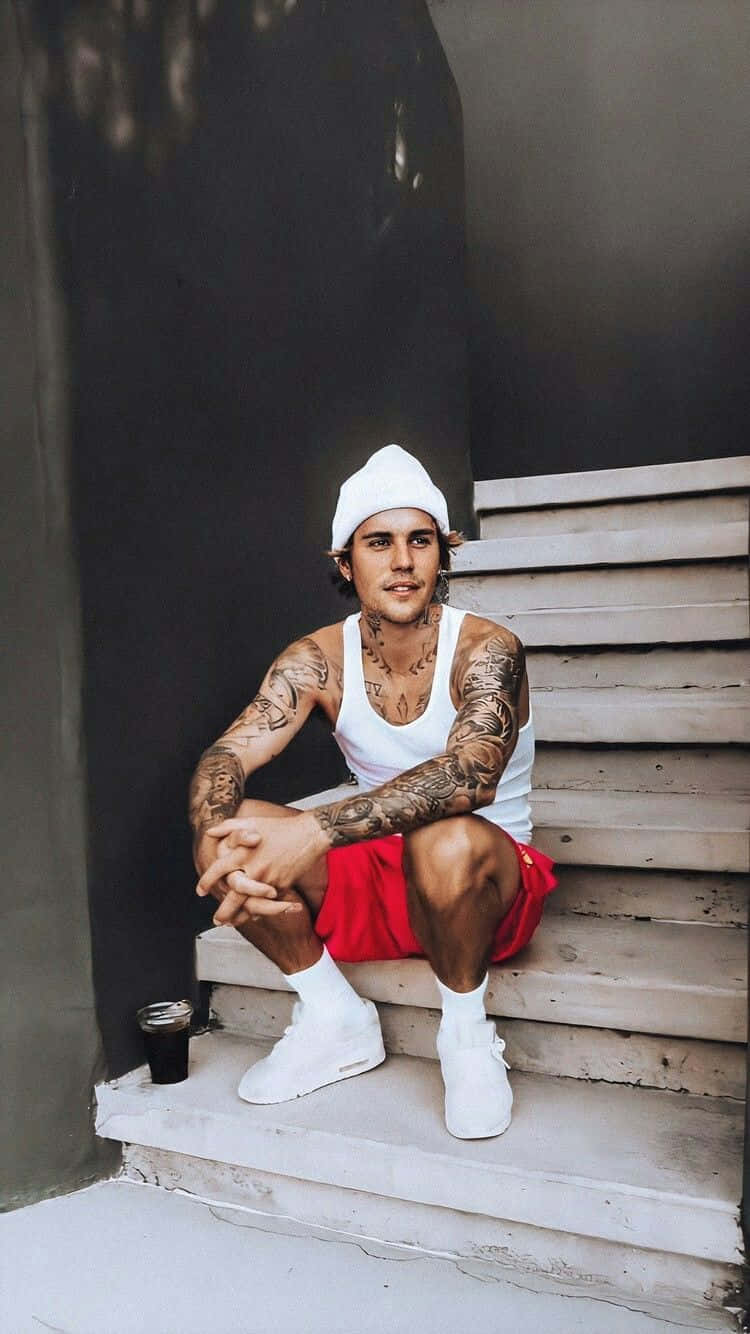 Justin Bieber 2021 桌布 壁紙 — 一個有刺青的男人，戴著白色毛帽和紅色短褲，隨意地坐在風化的混凝土階梯上，拿著飲料。