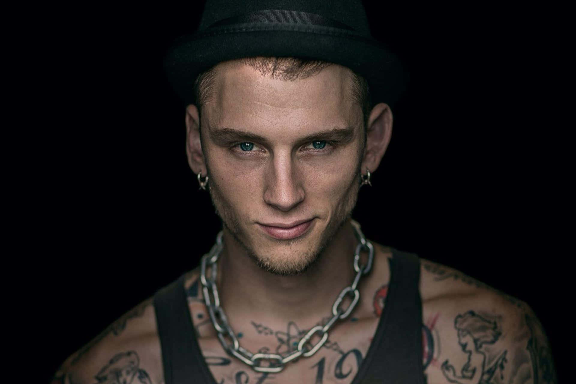 mgk 桌布 壁紙 — 一個有刺青的男人，戴著黑色軟呢帽和鏈條項鍊，在黑暗背景下凝視著。