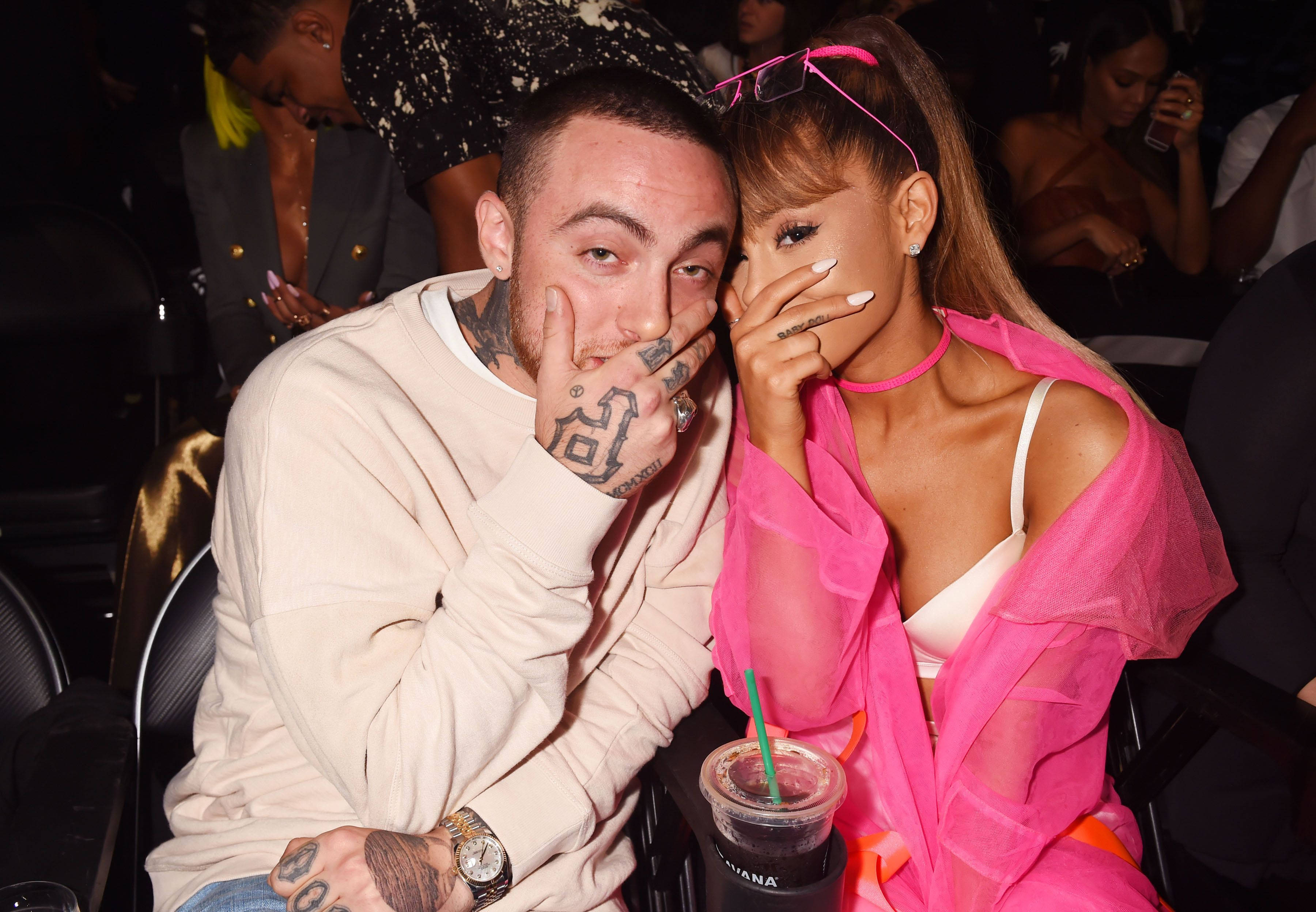 mac miller 4K 桌布 壁紙 — 一個有刺青的男人和一個穿粉紅色衣服的女人在擠滿人的活動中坐著，頑皮地遮住臉。