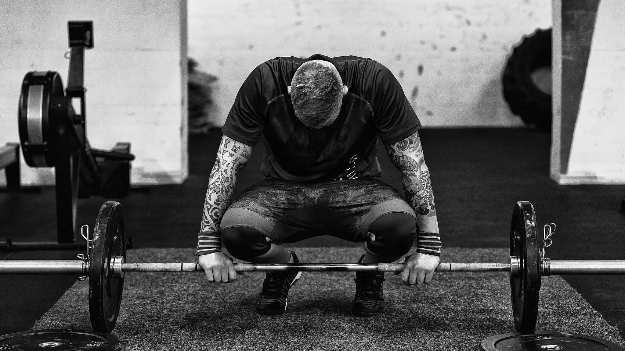 Fitness 桌布 壁紙 — 一個有刺青的男人在健身房中，黑白畫面下，蹲在一個裝有槓片的槓鈴上，神情專注。