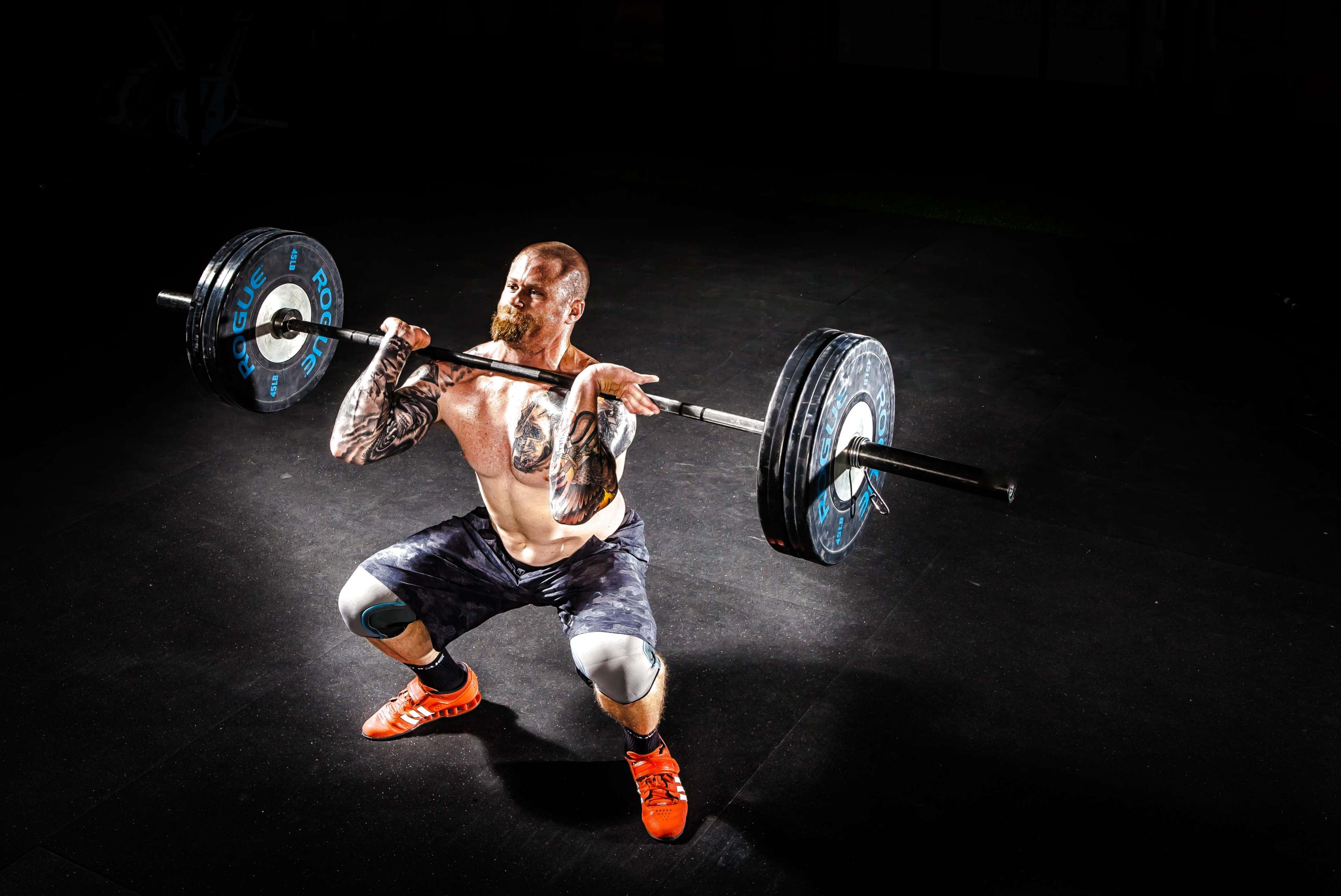 槓鈴 桌布 壁紙 — 一個有刺青的男人在健身房內，在深色背景下，專注地背負著沉重的槓鈴進行深蹲。