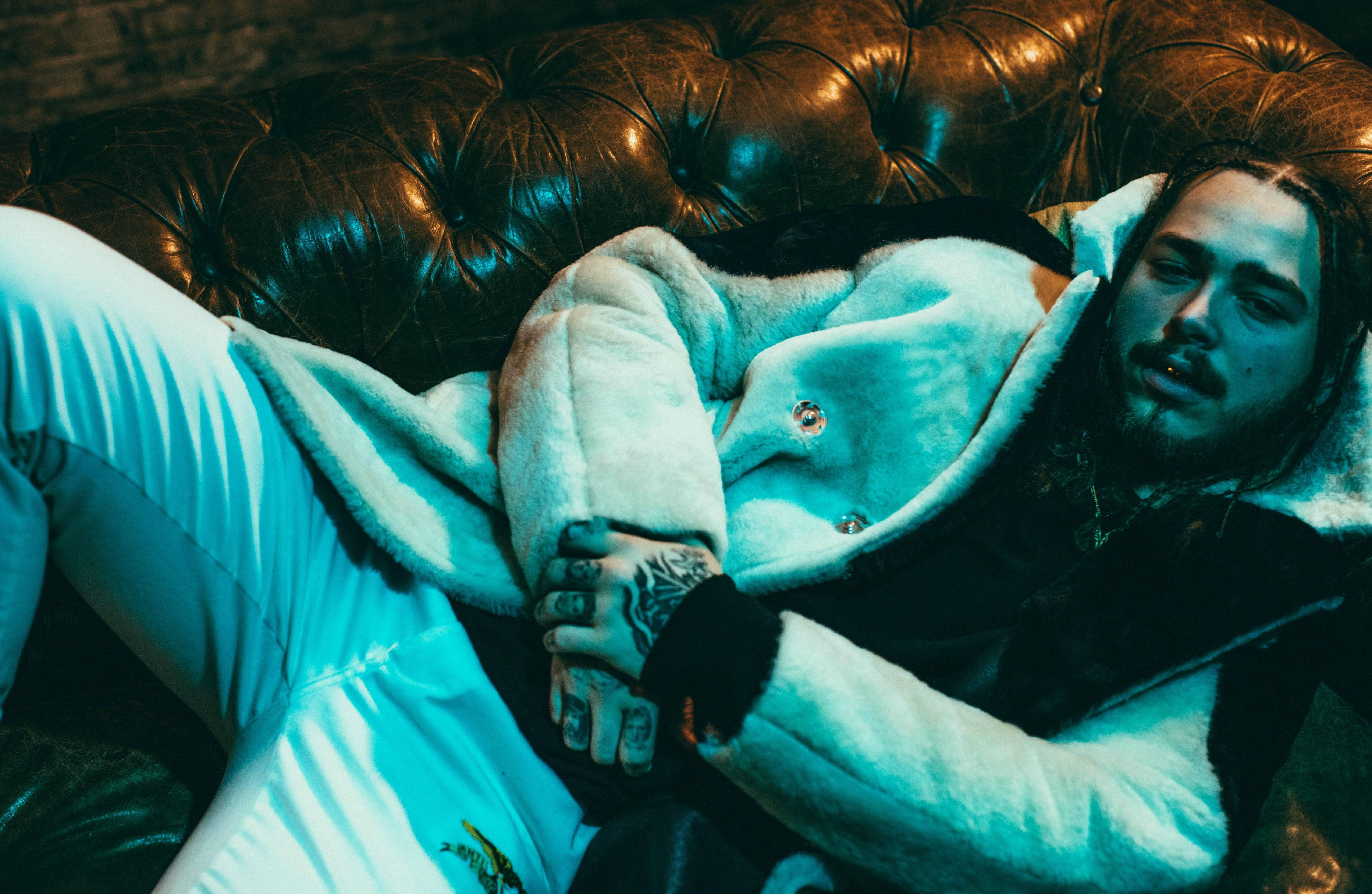 Post Malone 桌布 壁紙 — 一個有刺青的男人在冷藍燈光下，穿著毛茸茸的白色外套，靠在一張簇絨皮革沙發上休息。
