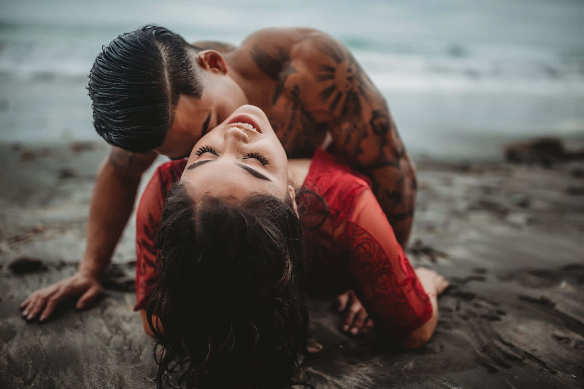 Couple Kissing 桌布 壁紙 — 一個有刺青的男人在濕漉漉的岩石海灘上俯身靠近一個穿著紅色連衣裙微笑的女人，身後波浪拍打。