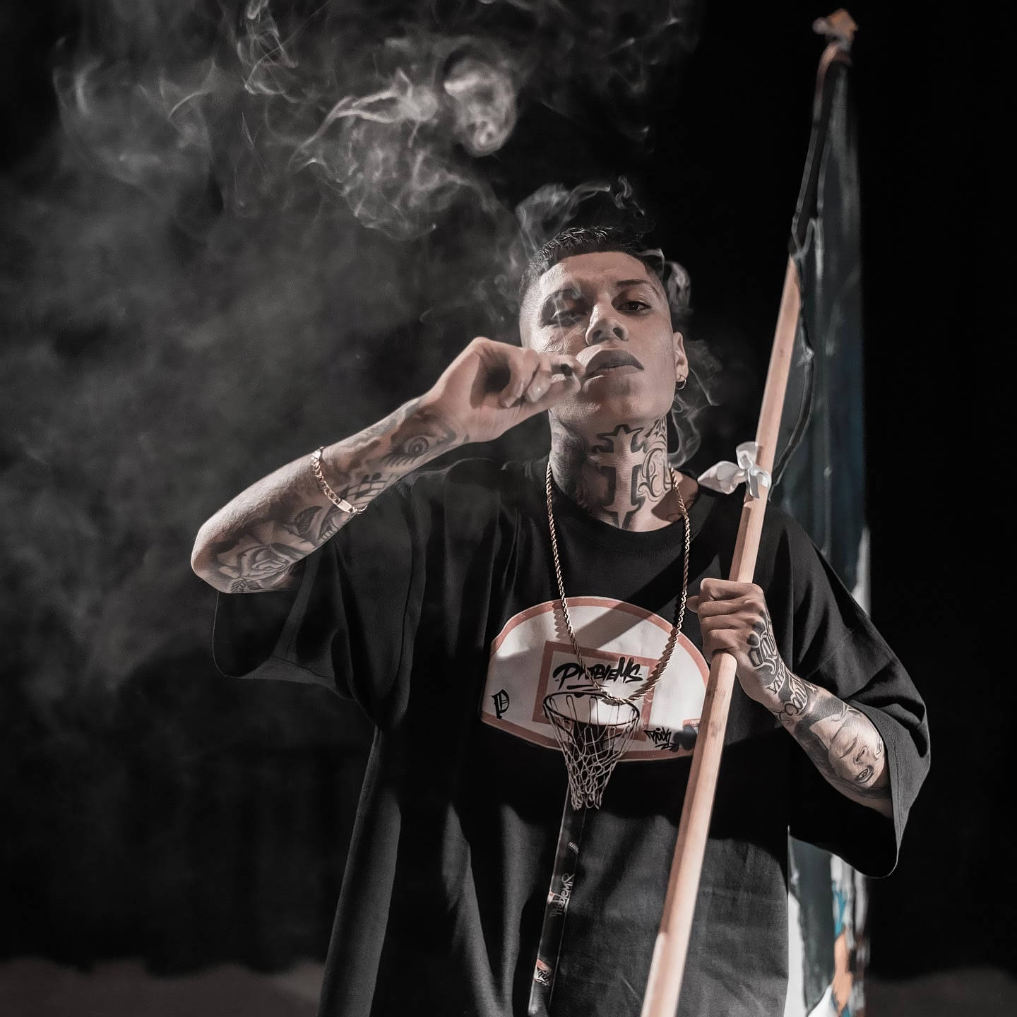 Fe 桌布 壁紙 — 一個有刺青的男人在煙霧瀰漫的黑暗背景下，一邊抽煙，一邊拿著旗幟和籃球框吊飾。