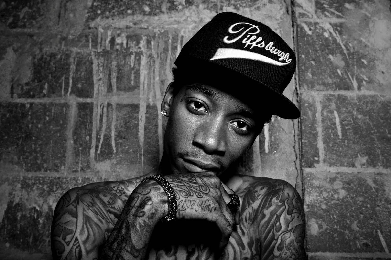 wiz khalifa 桌布 壁紙 — 一個有刺青的男人戴著 'Piff's laugh' 帽子，單手托腮靠在風化的磚牆上，黑白風格。