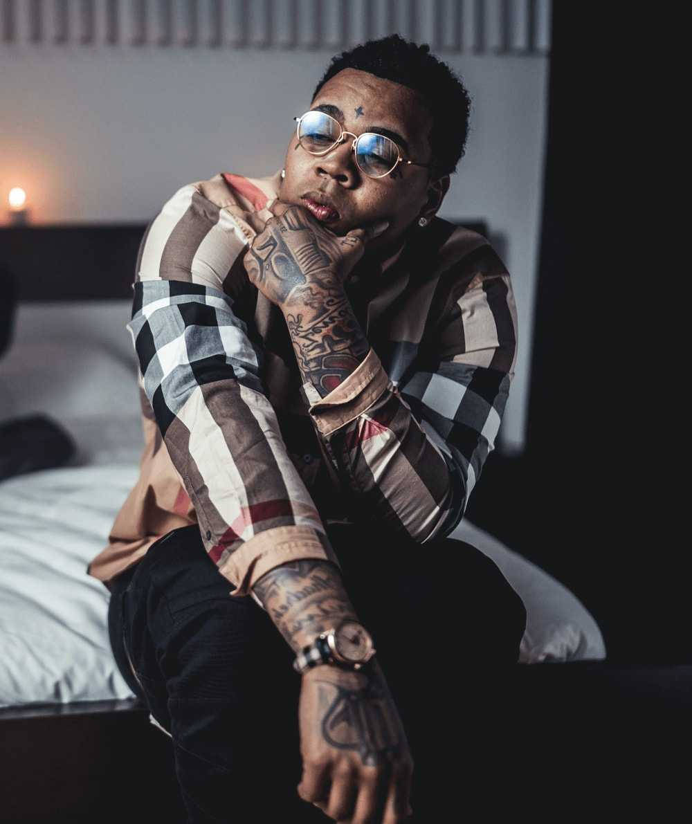 Kevin Gates 桌布 壁紙 — 一個有刺青的男人戴著圓形太陽眼鏡，在燭光氛圍下若有所思地坐在昏暗房間的床上。