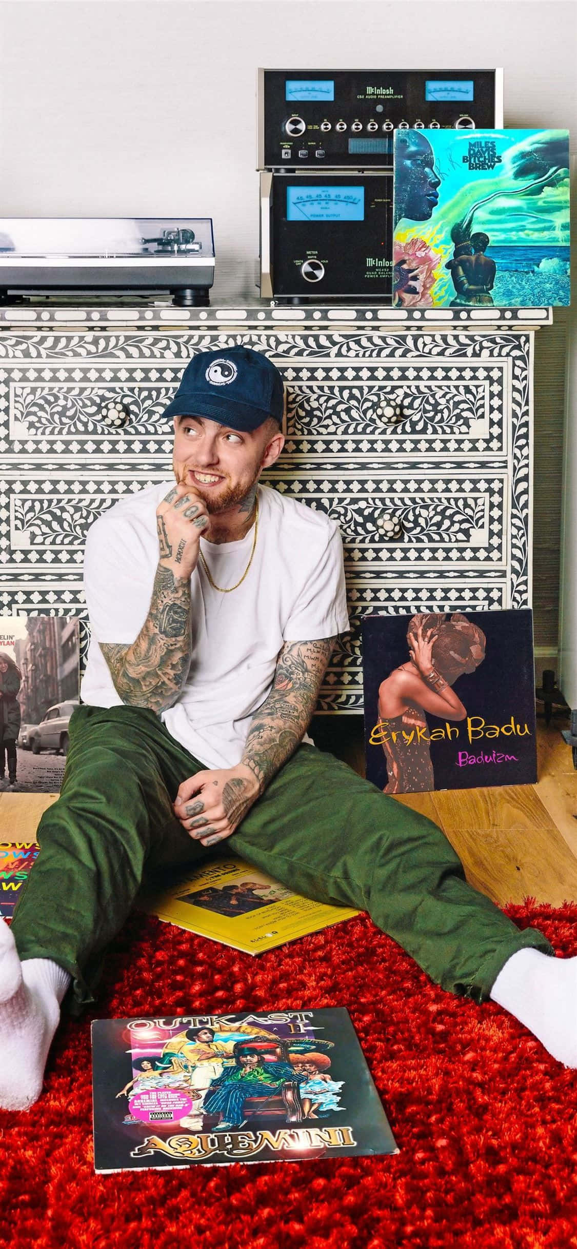 Hip Hop Album Covers 桌布 壁紙 — 一個有刺青的男人戴著帽子，盤腿坐在紅色長毛地毯上，周圍放滿黑膠唱片和復古音響系統。