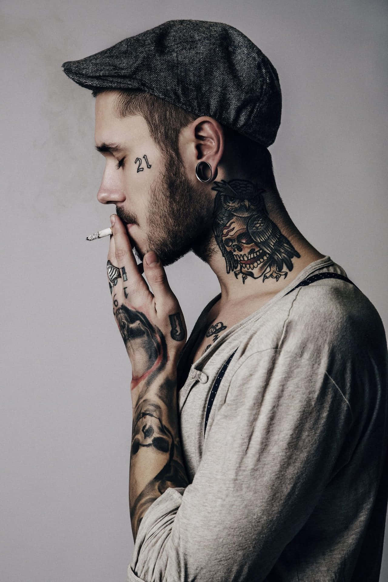 Tattoo Boy 桌布 壁紙 — 一個有刺青的男人戴著平頂帽，在純灰色背景前抽煙，臉上和頸部有明顯的刺青。