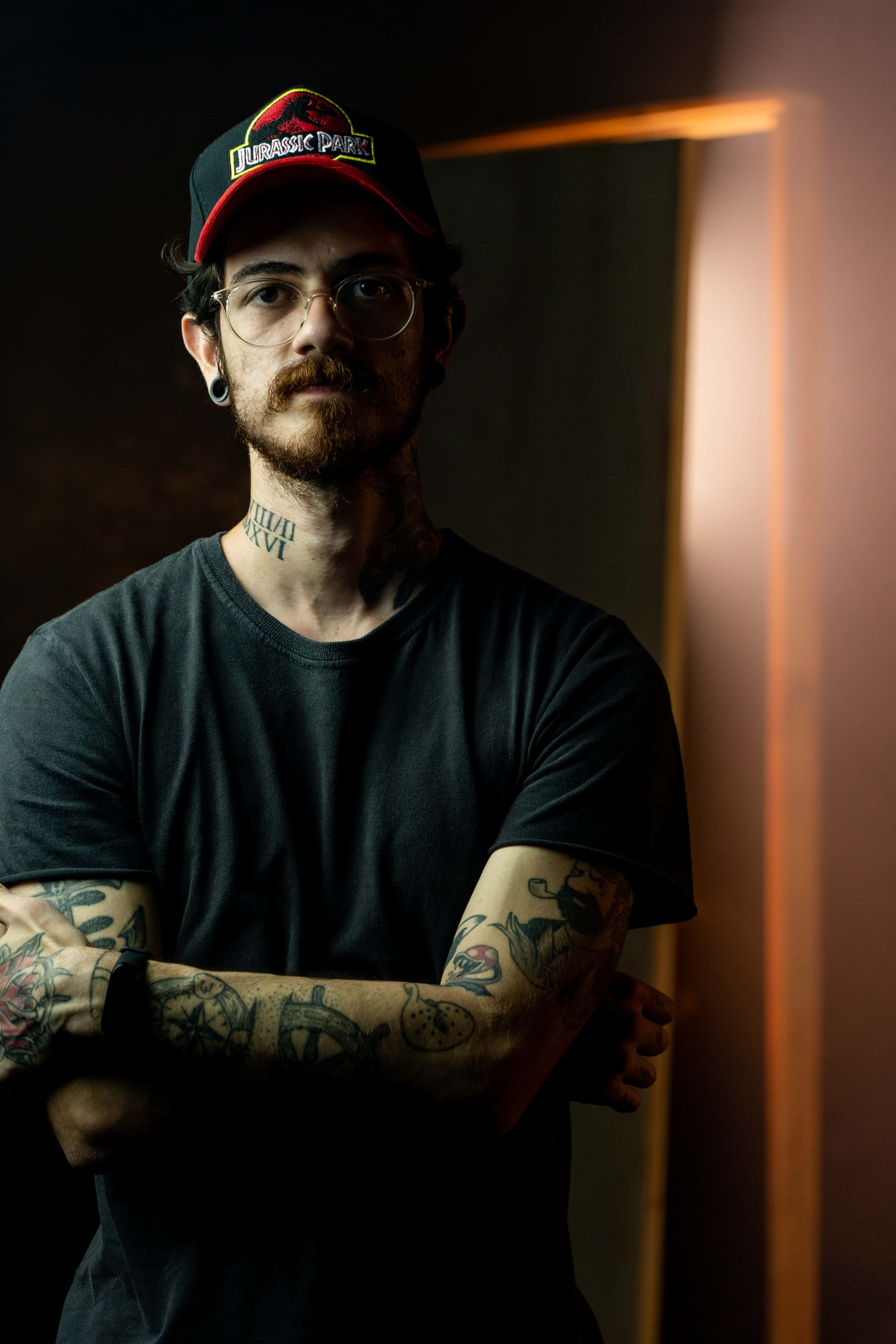 Tattoo 桌布 壁紙 — 一個有刺青的男人戴著眼鏡和Jurassic Park帽，在昏暗的房間裡自信地站著，背景有溫暖的燈光。
