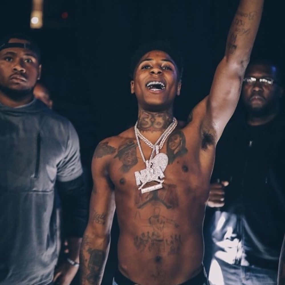 Youngboy 桌布 壁紙 — 一個有刺青的男人沒穿上衣戴著鑽石項鍊，在昏暗的房間裡高舉手臂，身後有兩個男人。
