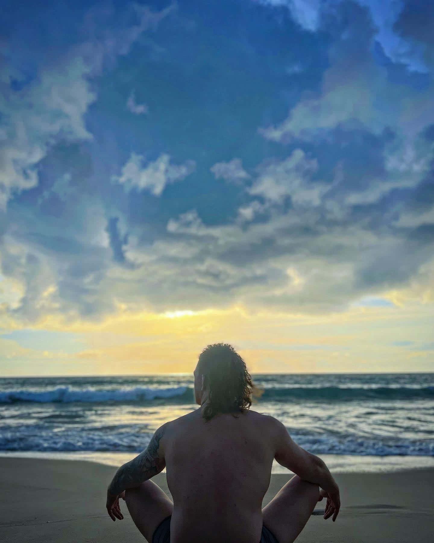 海灘 桌布 壁紙 — 一個有刺青的男人盤腿坐在沙灘上，凝視著海洋，金色的夕陽光線穿過戲劇性的藍色雲層。