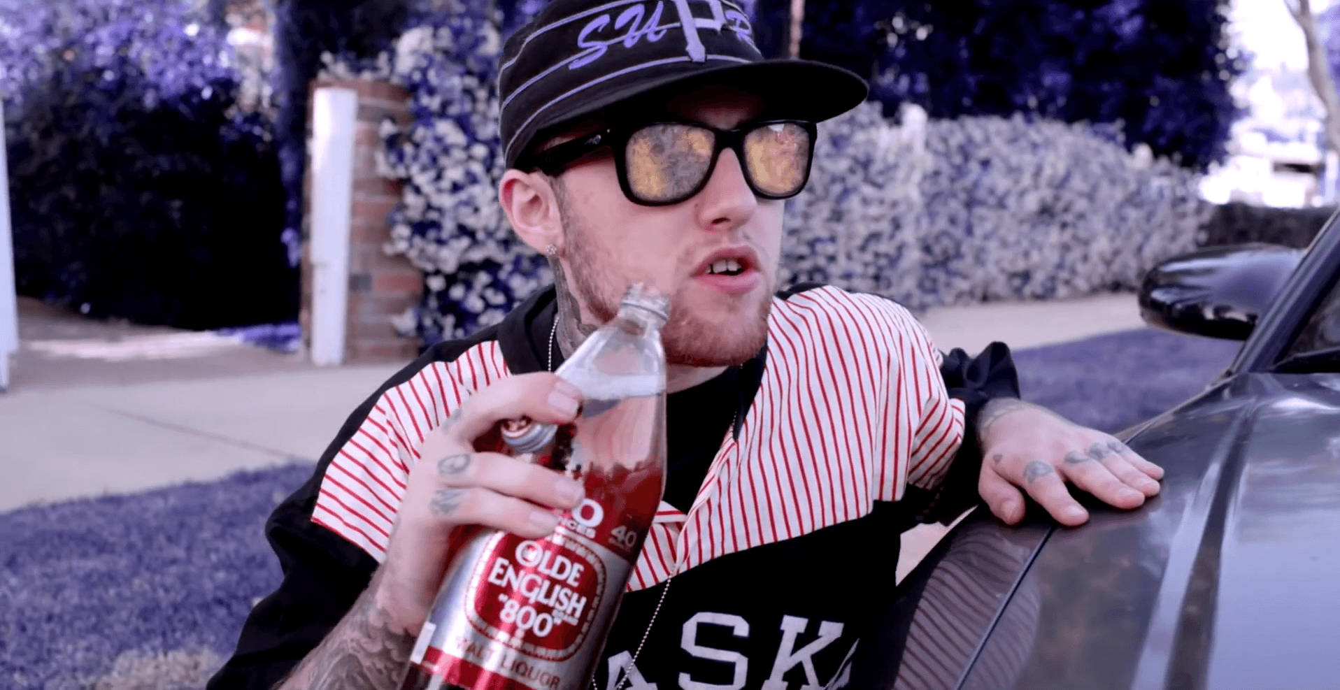 Mac Miller 桌布 壁紙 — 一個有刺青的男人穿著條紋襯衫和帽子，靠著車子喝一口Olde English 800的瓶子。