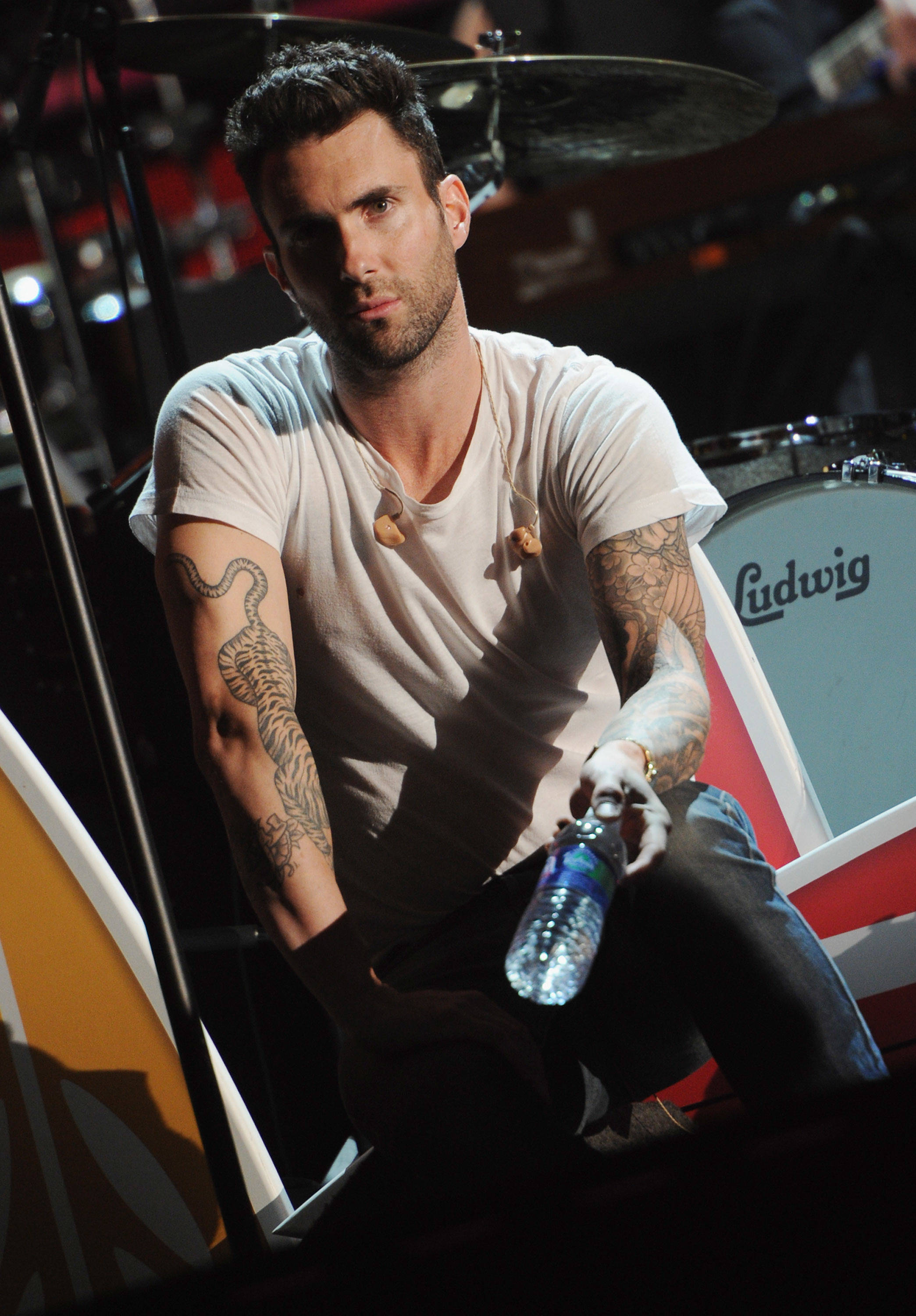 Adam Levine 桌布 壁紙 — 一個有刺青的男人穿著白色T恤，在舞台燈光下坐在Ludwig鼓組旁邊，拿著水瓶。