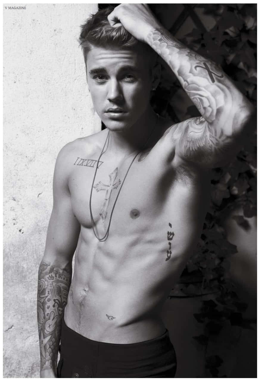 Justin Bieber 2015 桌布 壁紙 — 一個有刺青的男人赤裸上身，頭髮造型有型，靠著一面有紋理和常春藤的牆，手放在頭上，以單色調擺姿勢。