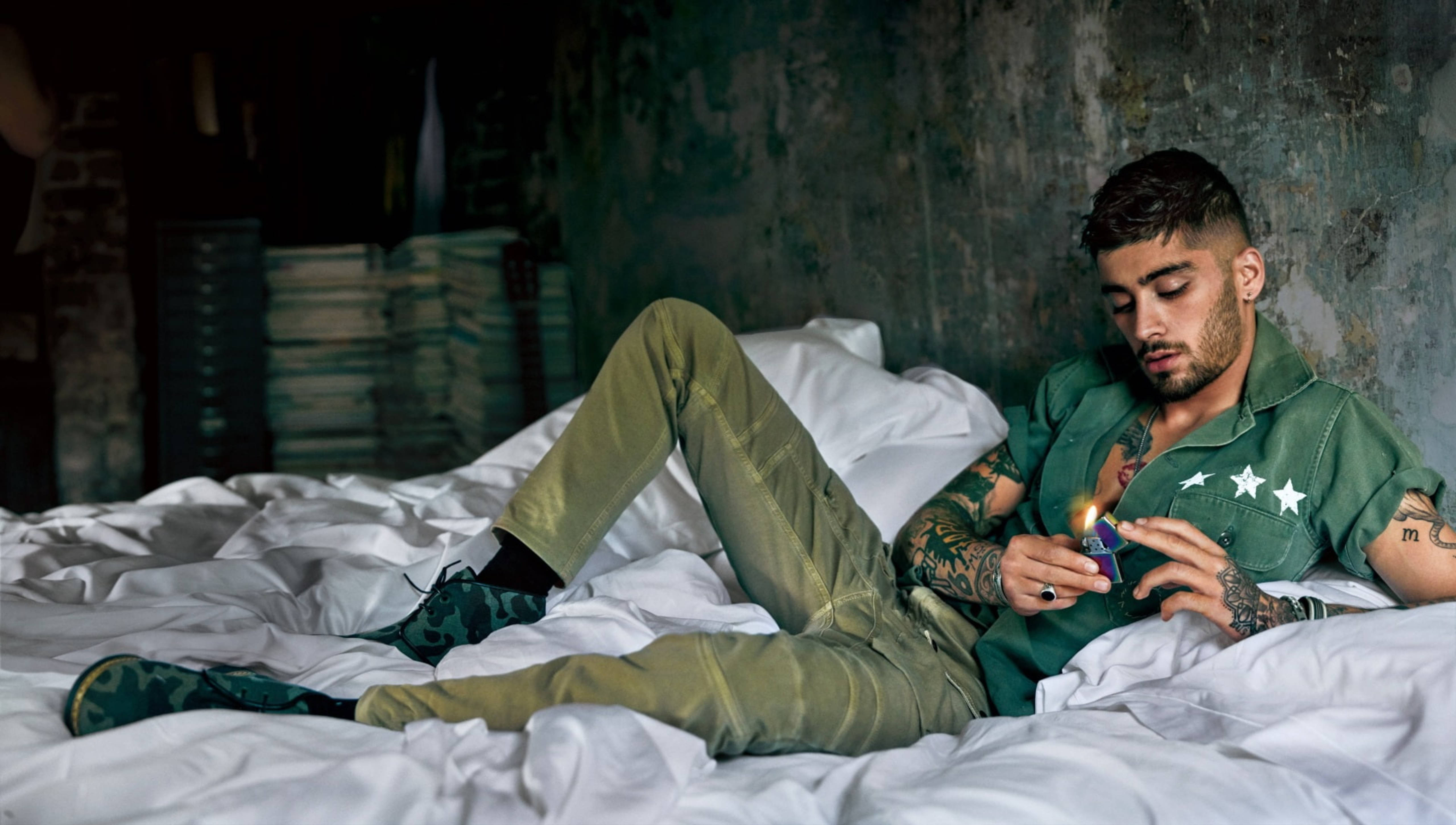 zayn 桌布 壁紙 — 一個有刺青的男人躺在昏暗房間裡一張凌亂的床上，專注地點燃打火機。