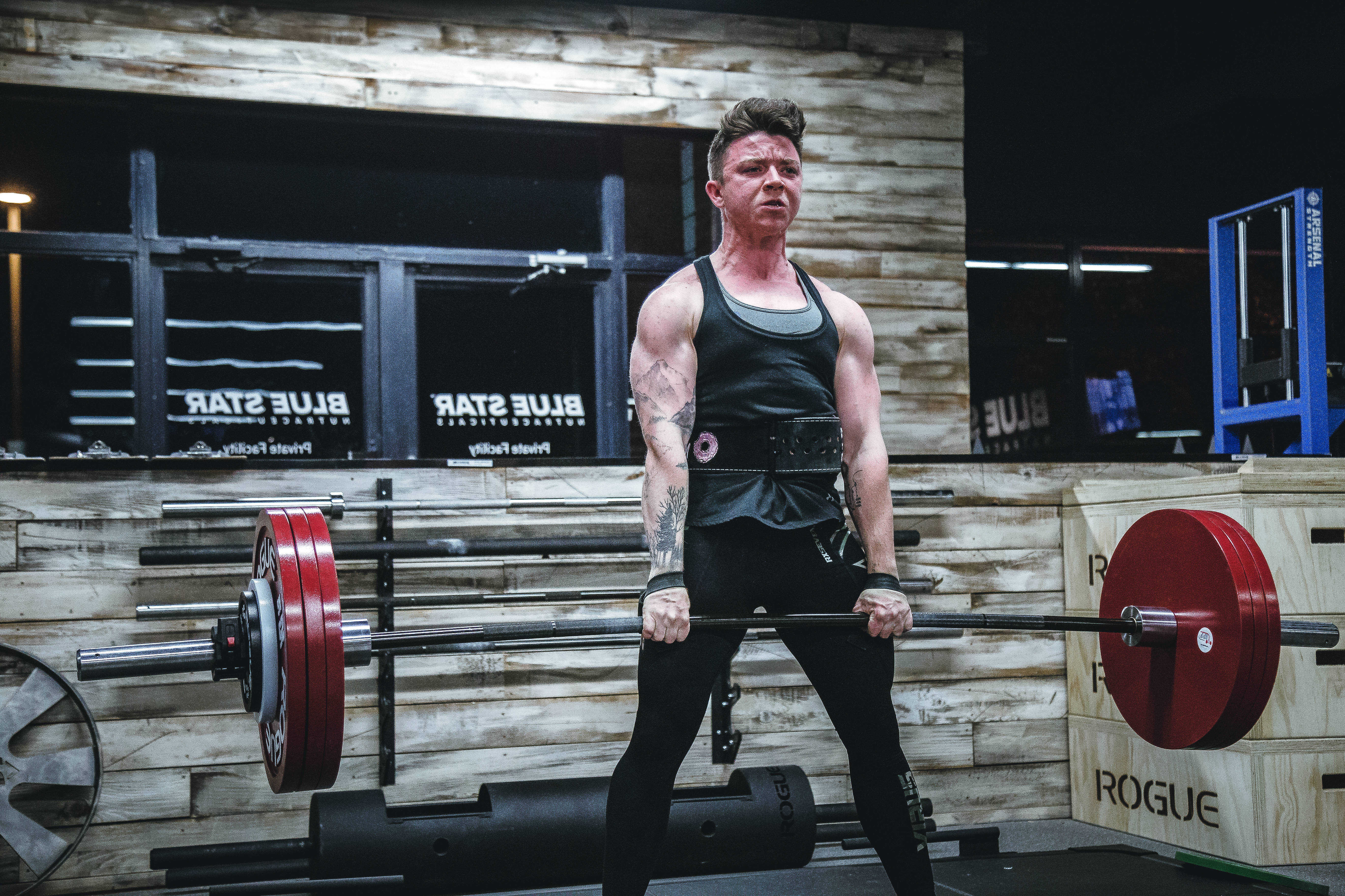 Weightlifting 桌布 壁紙 — 一個有刺青的肌肉男在一個有木牆和Rogue Fitness器材的鄉村風格健身房中，抓住一個裝有紅色槓鈴片的沉重槓鈴。