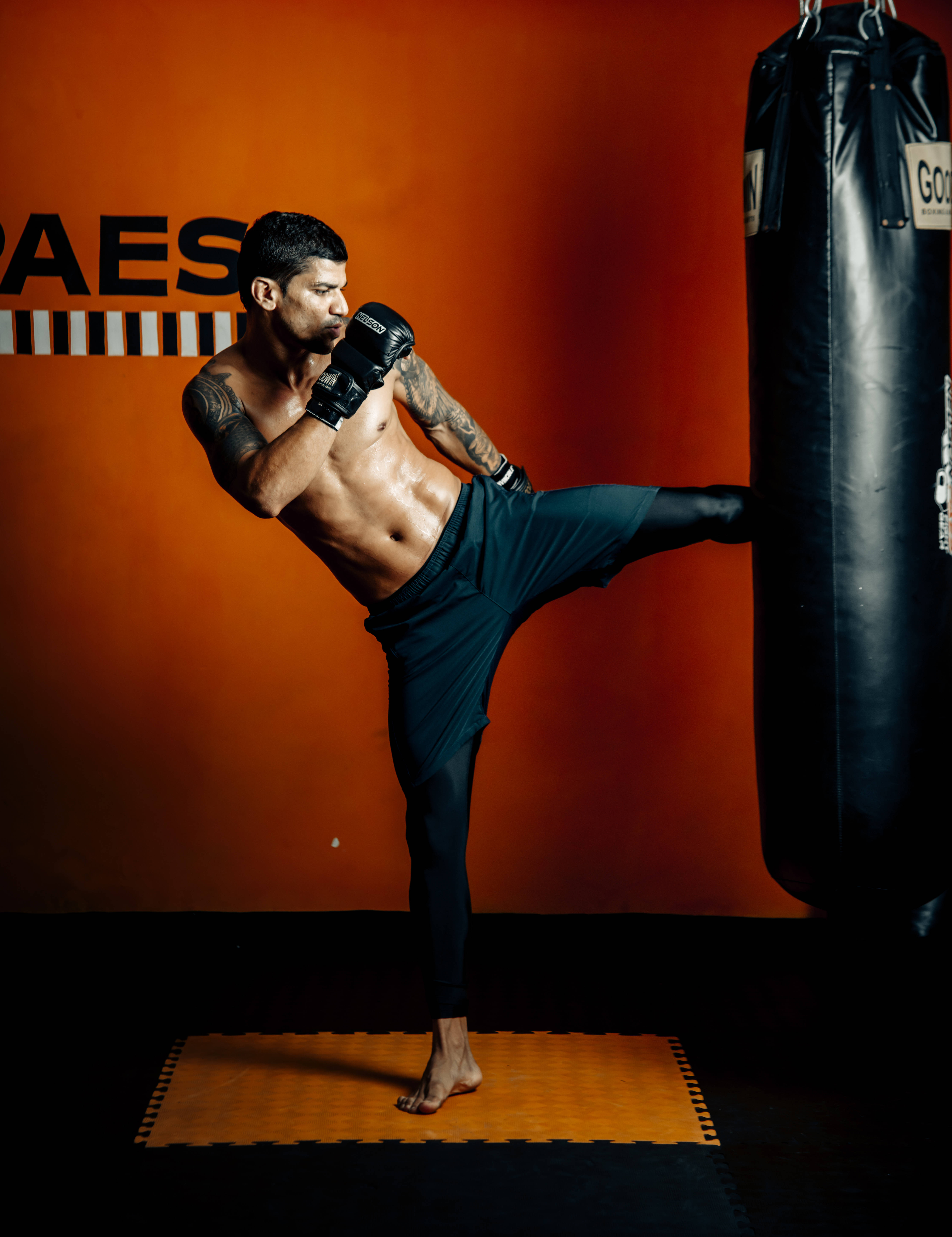 MMA 桌布 壁紙 — 一個有刺青的肌肉男在昏暗的橙色牆壁健身房中對重型沙袋進行高踢。