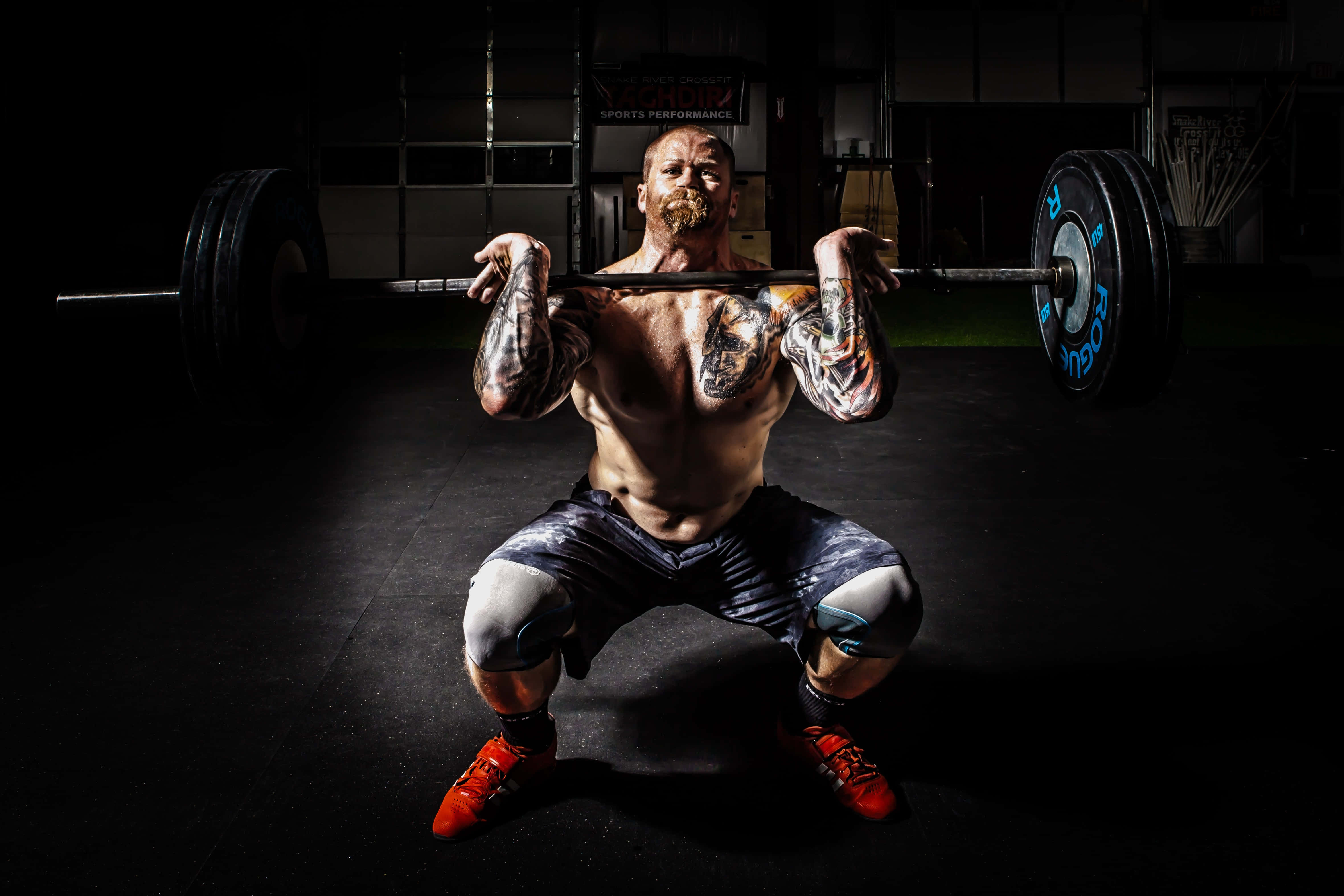 Barbell 桌布 壁紙 — 一個有刺青的肌肉男在燈光昏暗的健身房中，在戲劇性的聚光燈下進行槓鈴深蹲。