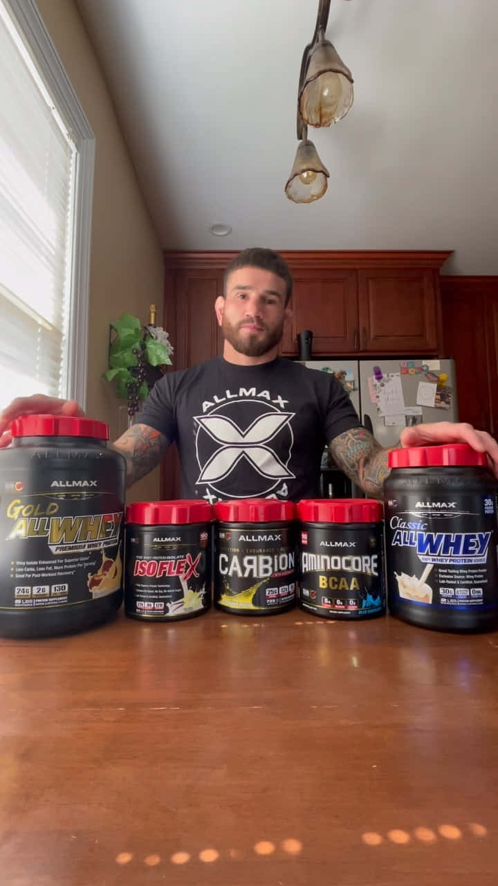 MMA 桌布 壁紙 — 一個有刺青的肌肉男站在廚房檯面後，展示五個大型Allmax Nutrition補充劑容器。