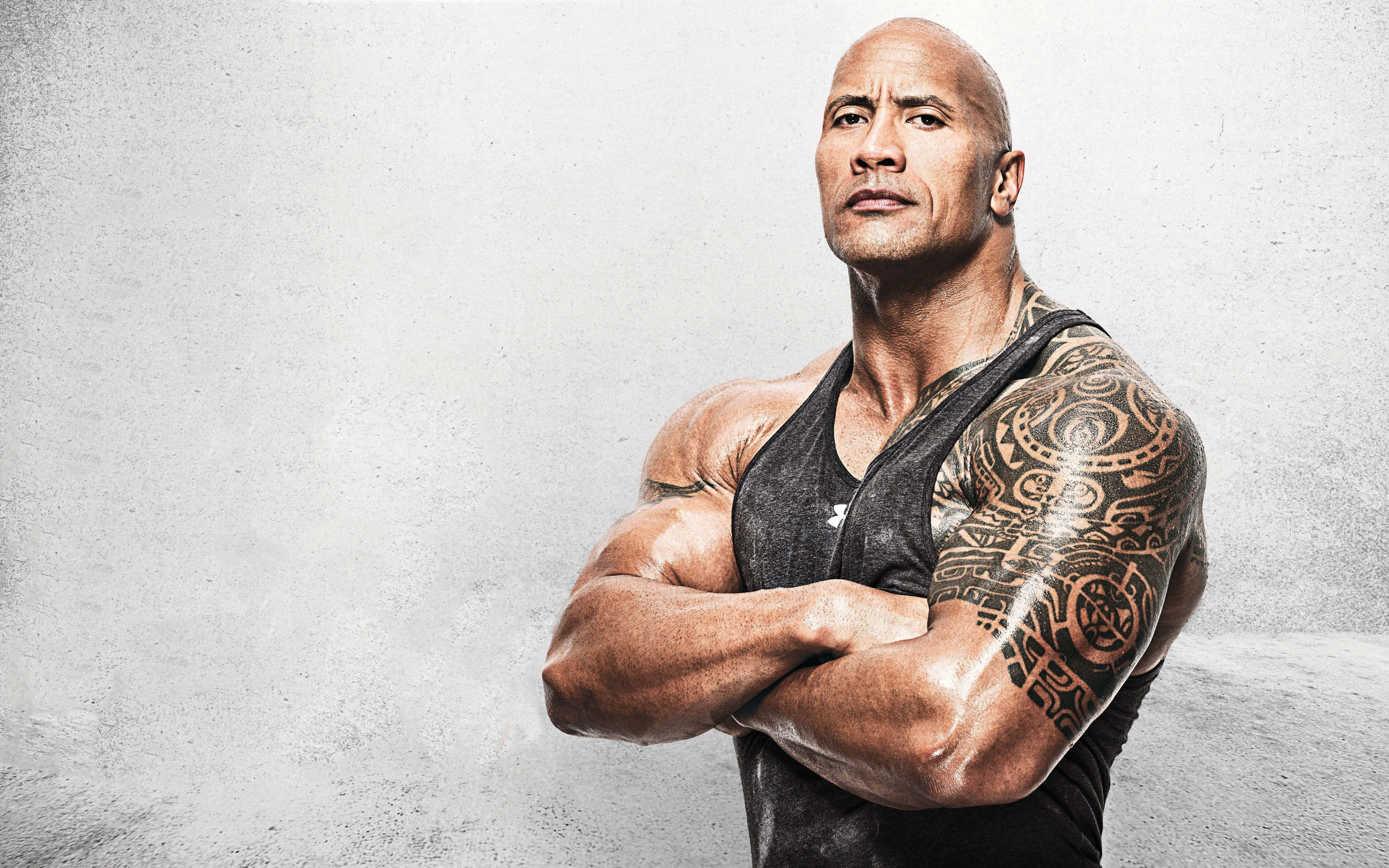 The Rock 桌布 壁紙 — 一個有刺青的肌肉男自信地交叉雙臂，靠著紋理灰色牆壁站立。