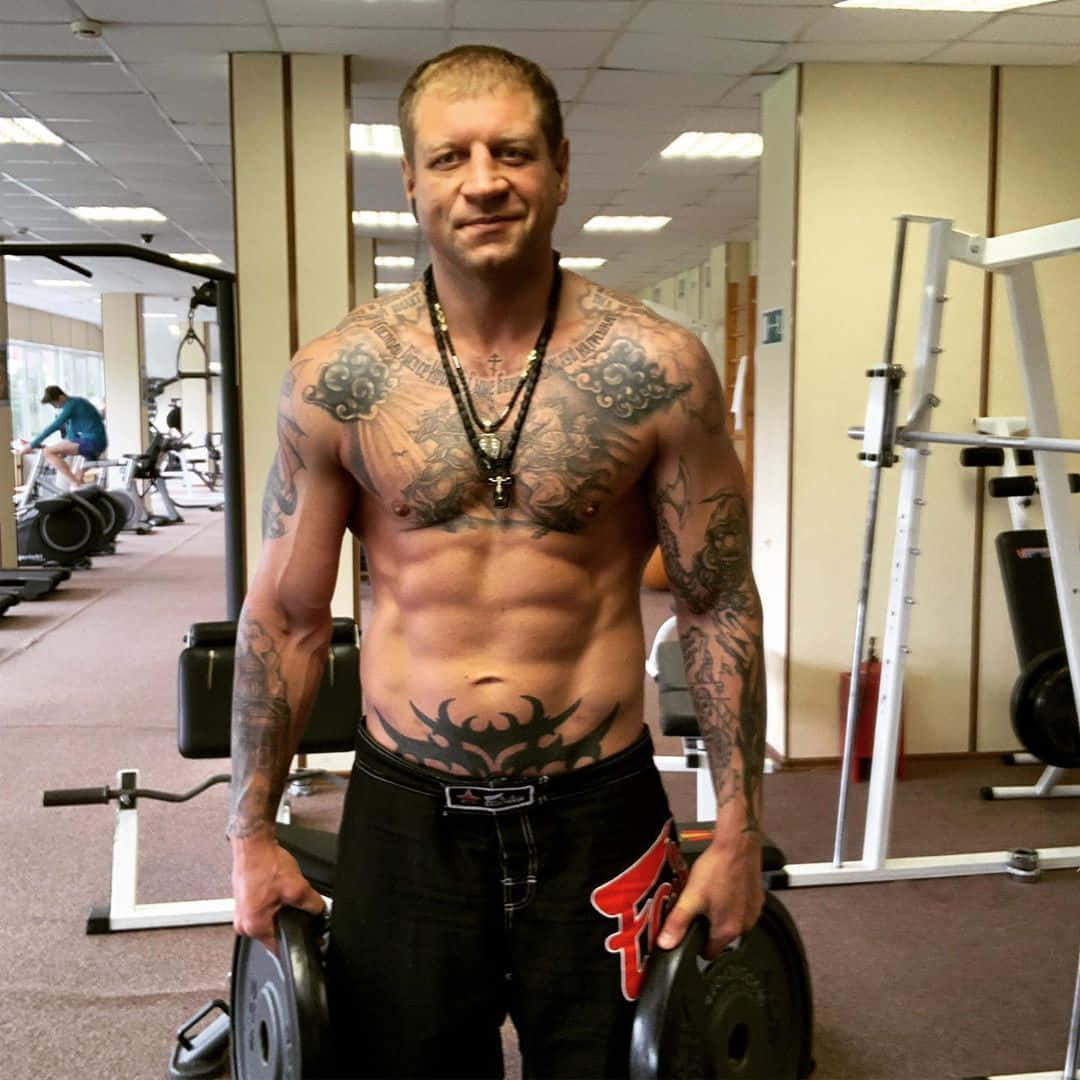 MMA 桌布 壁紙 — 一個有刺青的肌肉男赤裸上身站在健身房，自信地微笑著拿著槓片。