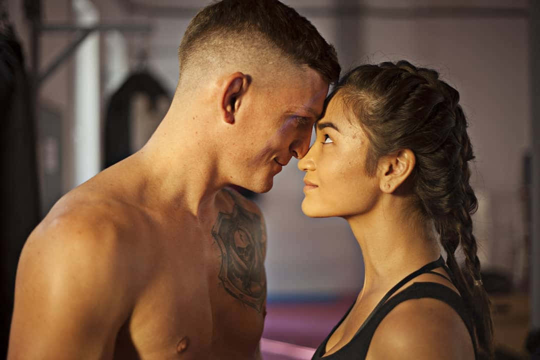 UFC 桌布 壁紙 — 一個有刺青的赤膊男人和一個穿著黑色背心的女人在柔和燈光的健身房裡額頭相觸，分享親密時刻。