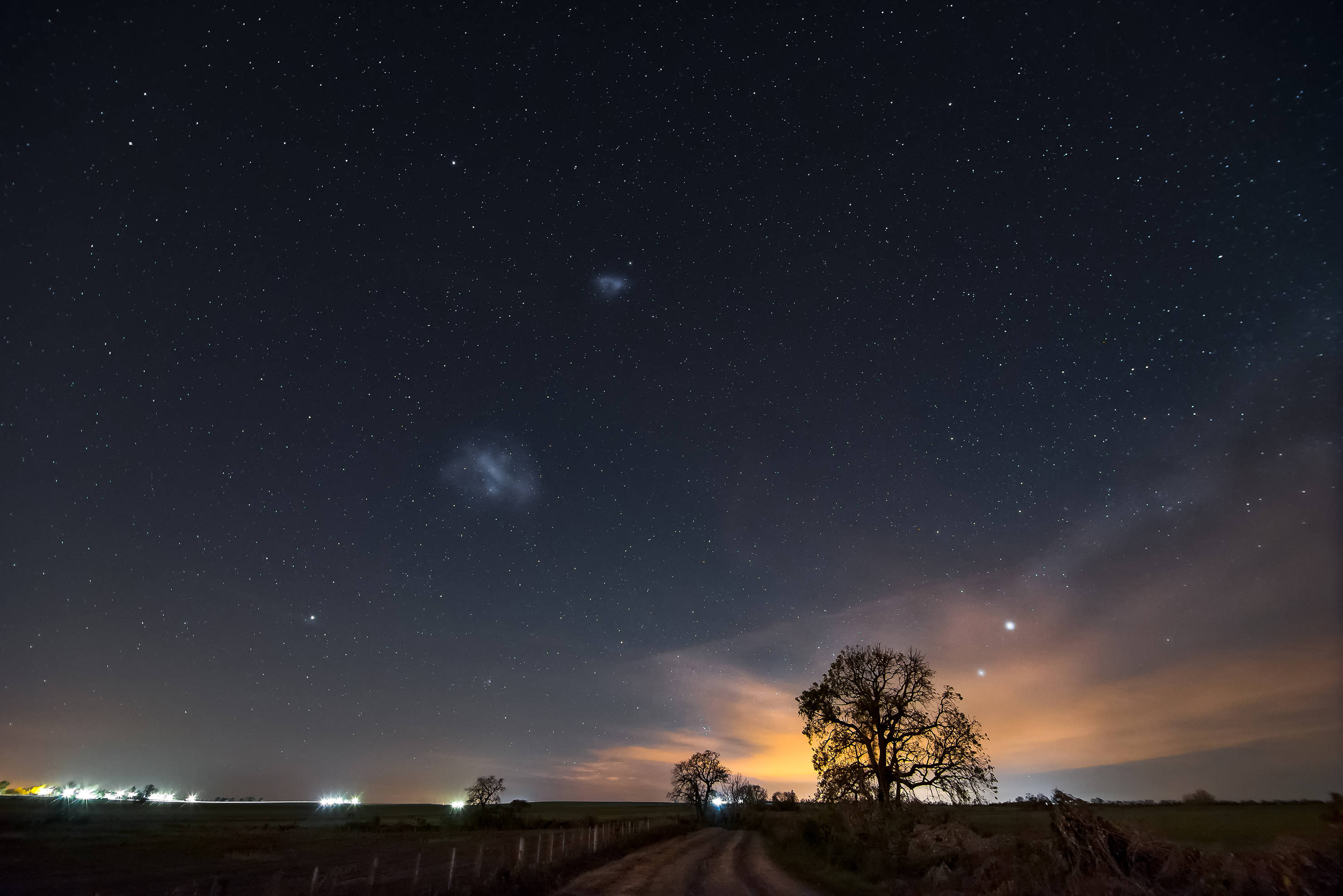 Starry 桌布 壁紙 — 一個有可見銀河系的星空夜晚，拱形在鄉村泥路上，兩旁是剪影樹木和遠處燈光。