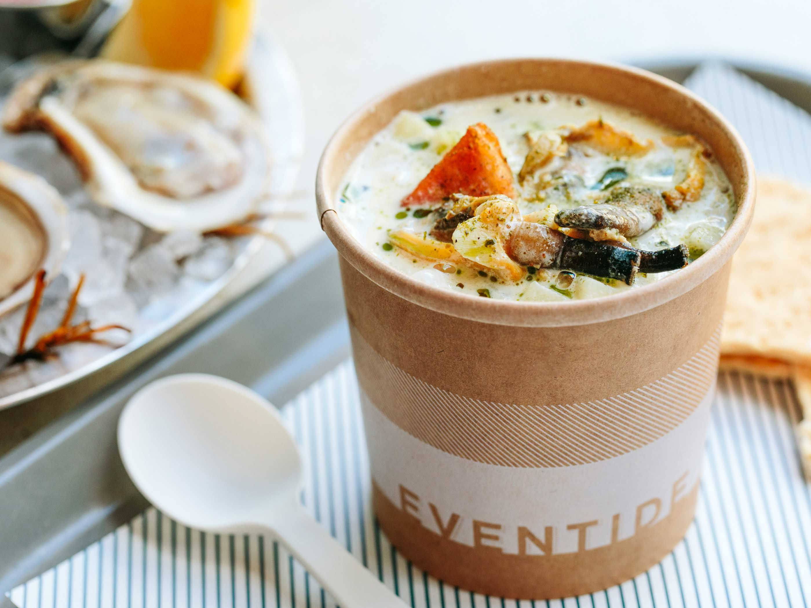 Chowder 桌布 壁紙 — 一個有品牌標誌的紙杯裝著奶油海鮮濃湯，旁邊放著冰上的新鮮生蠔和白色湯匙，在條紋托盤上。